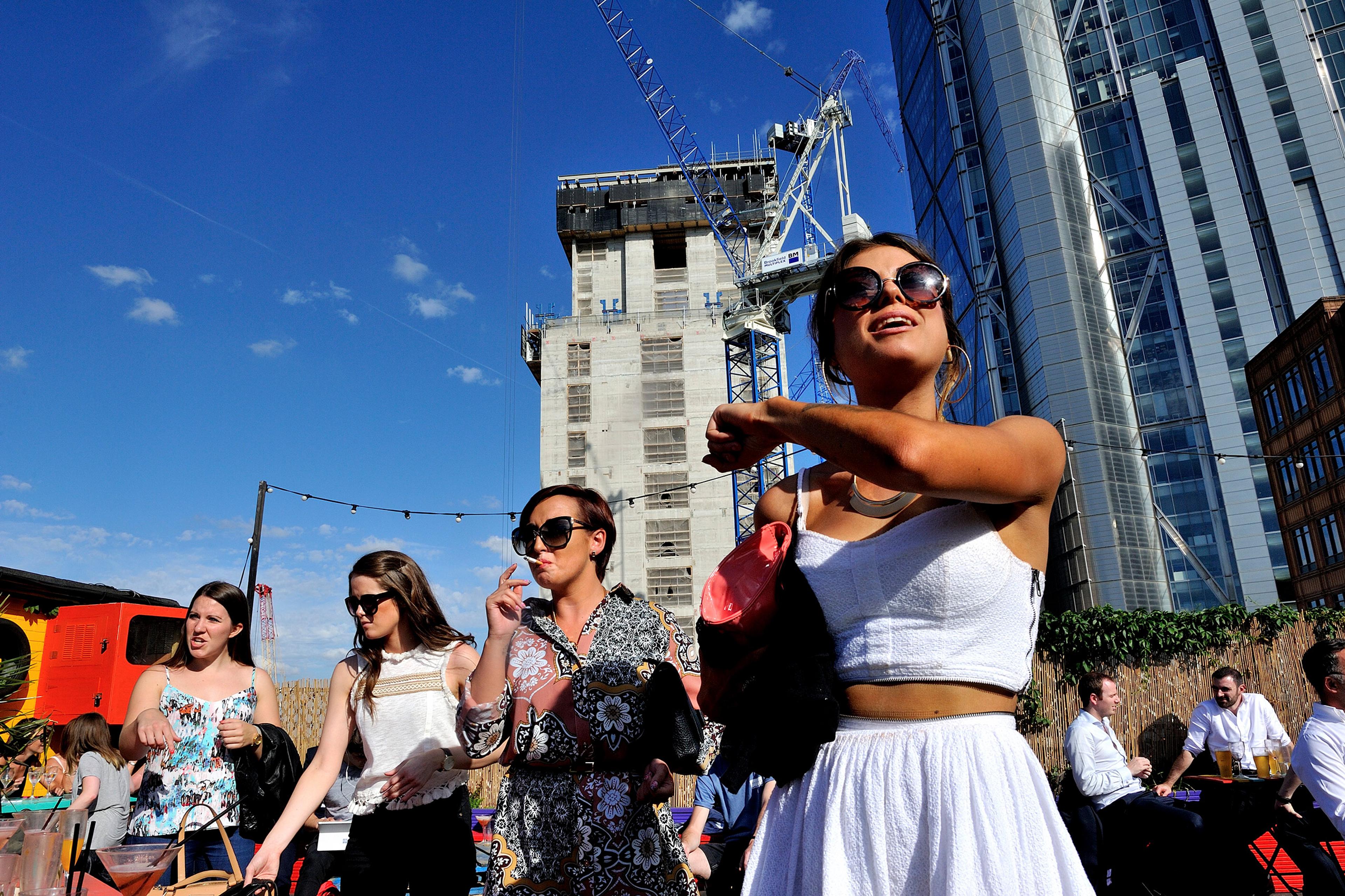 Four women outside wearing summer outfits with skyscrapers and a construction crane in the background; one woman is shown smoking.