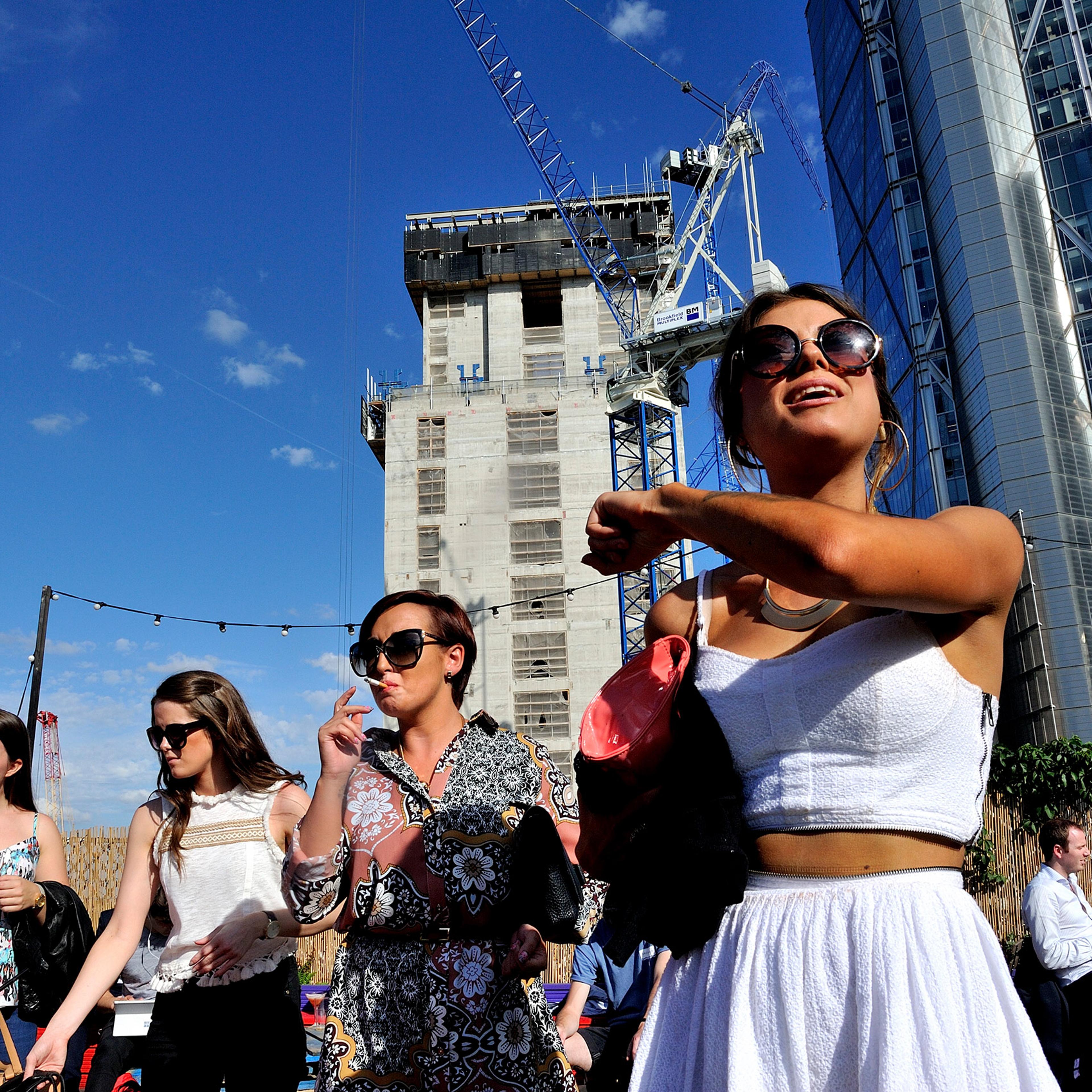 Four women outside wearing summer outfits with skyscrapers and a construction crane in the background; one woman is shown smoking.