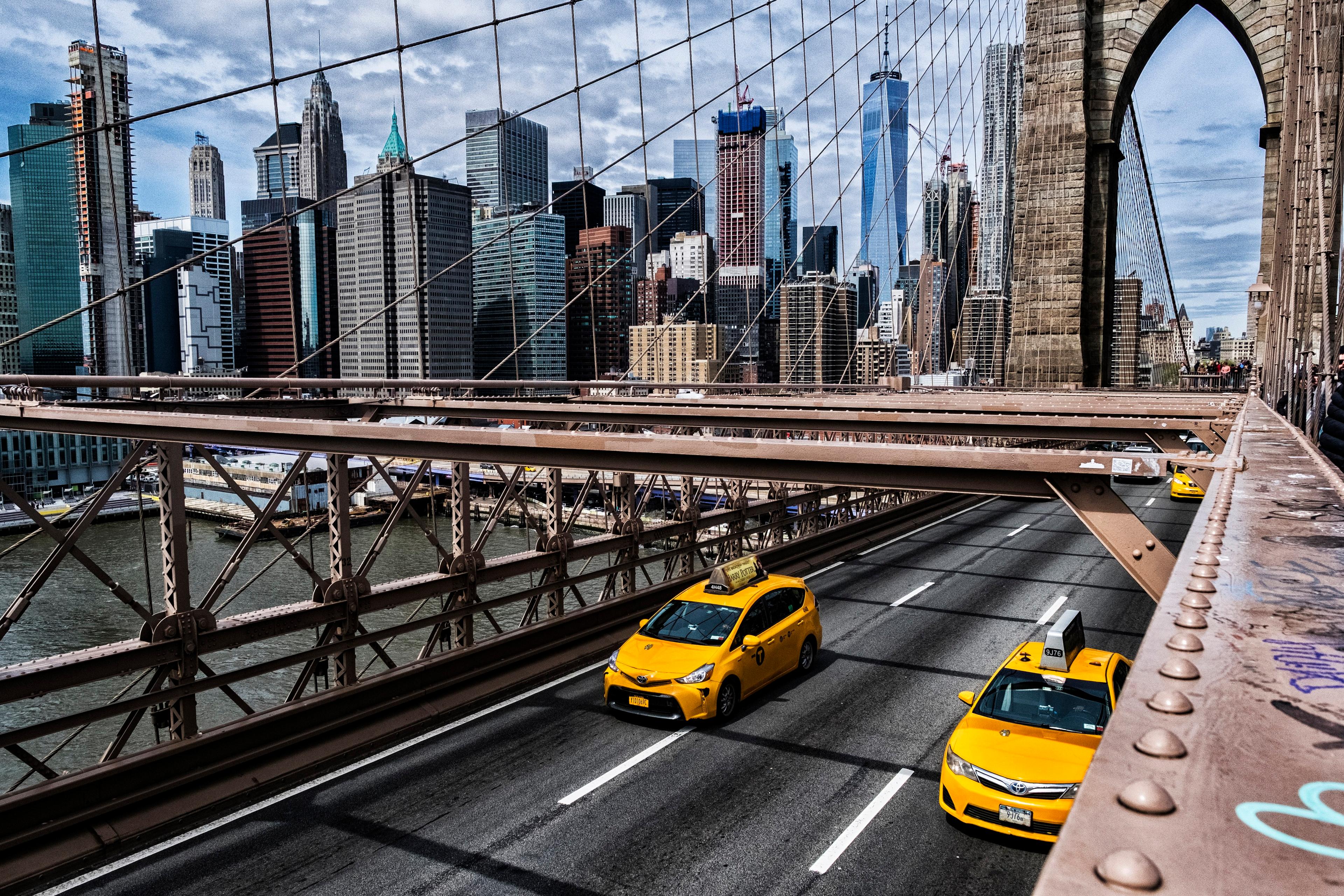 Photo of yellow taxis on Brooklyn Bridge with New York City skyline in the background on a cloudy day.
