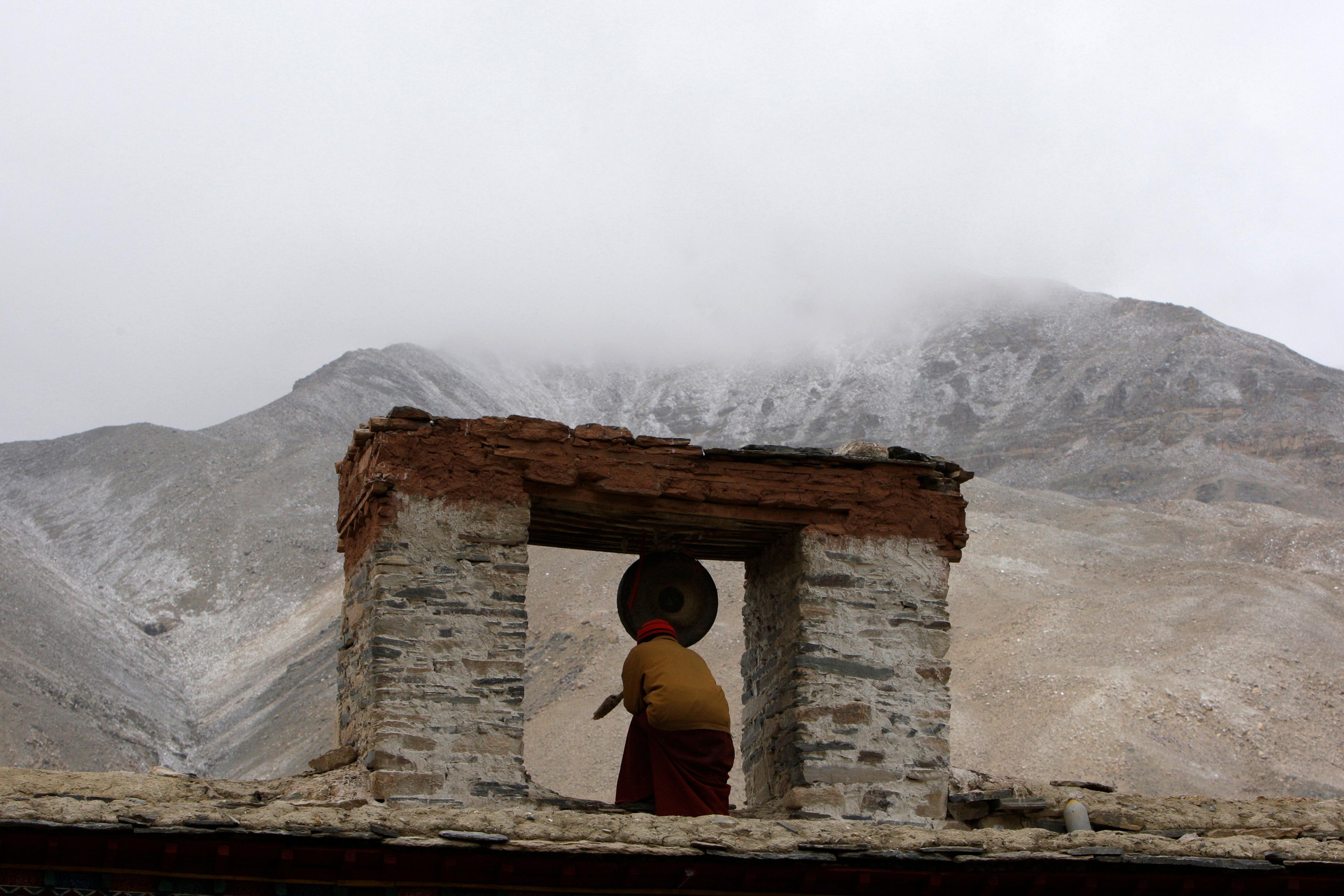 Photo of a monk striking a large gong in an open stone structure with misty mountains in the background.