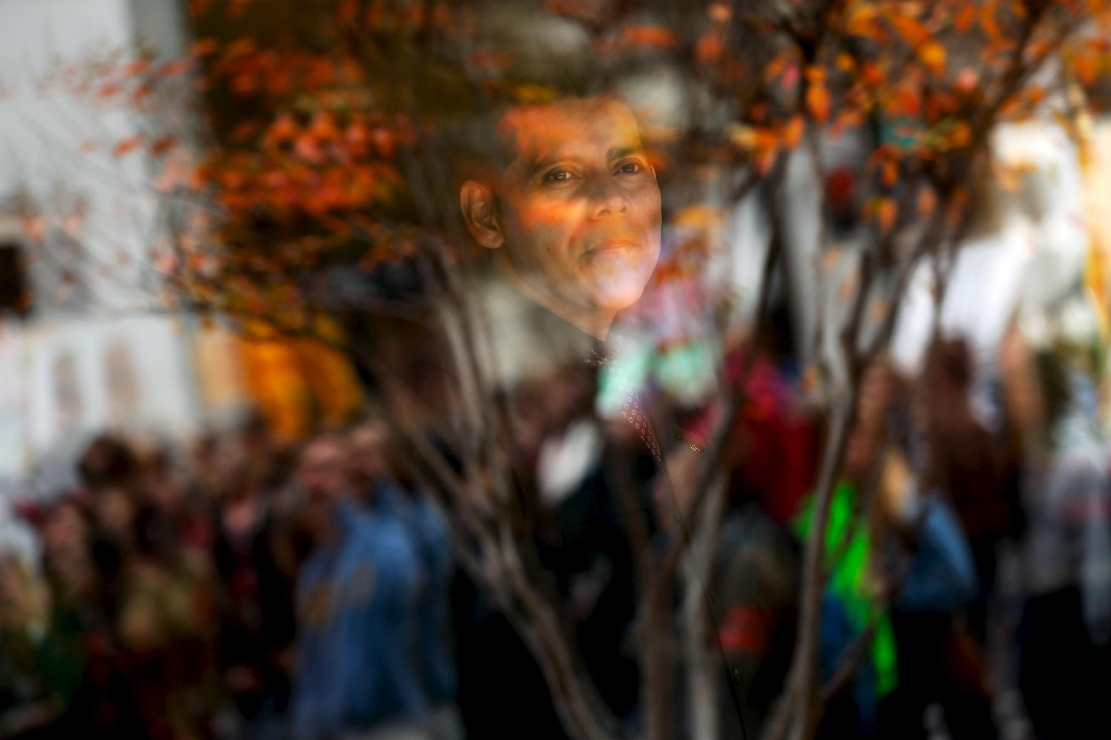 Photo showing a man’s reflection in glass with blurred people and tree branches in the foreground.