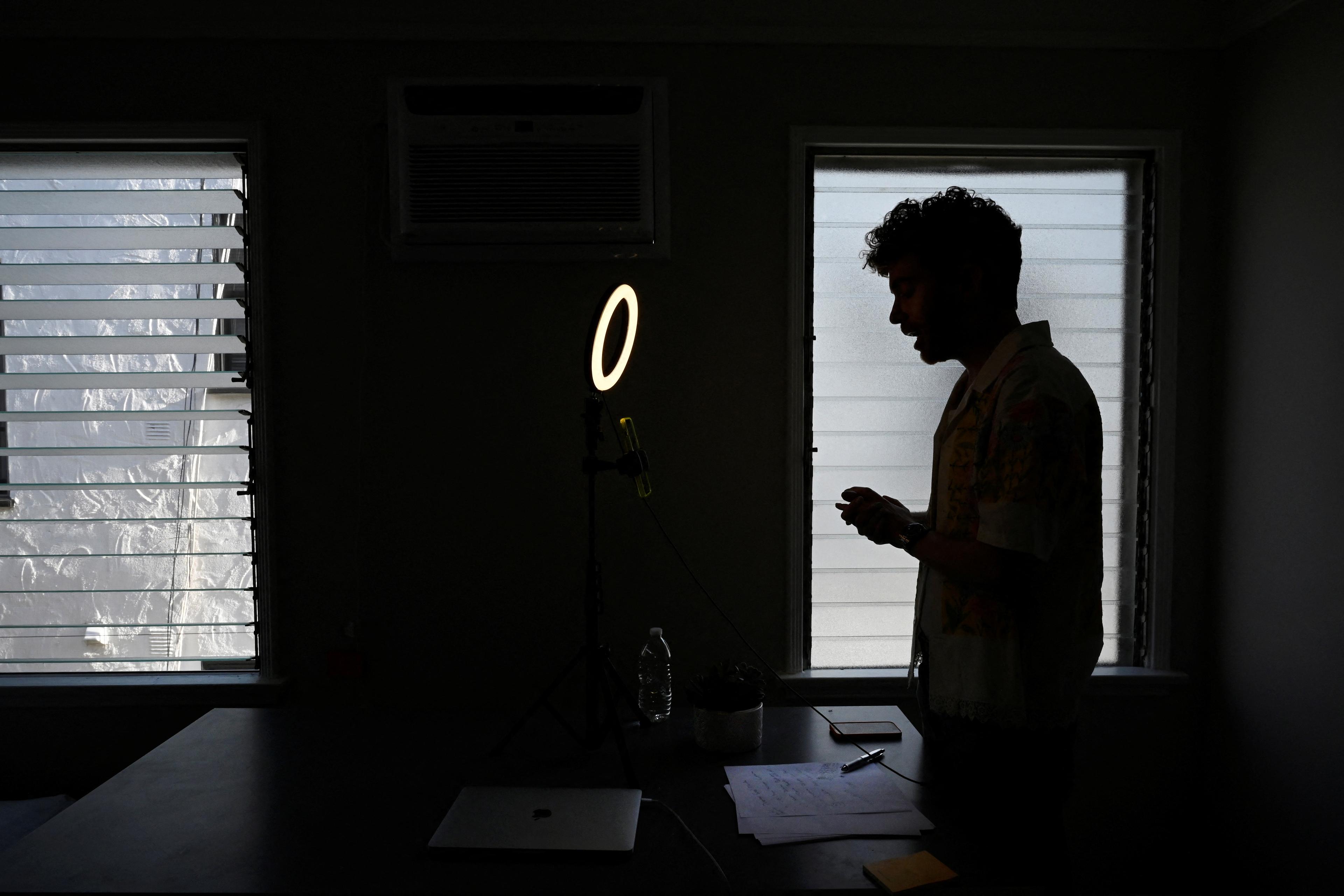Silhouette of a man by a window in a dim room with a ring light laptop and papers on the table, photo.