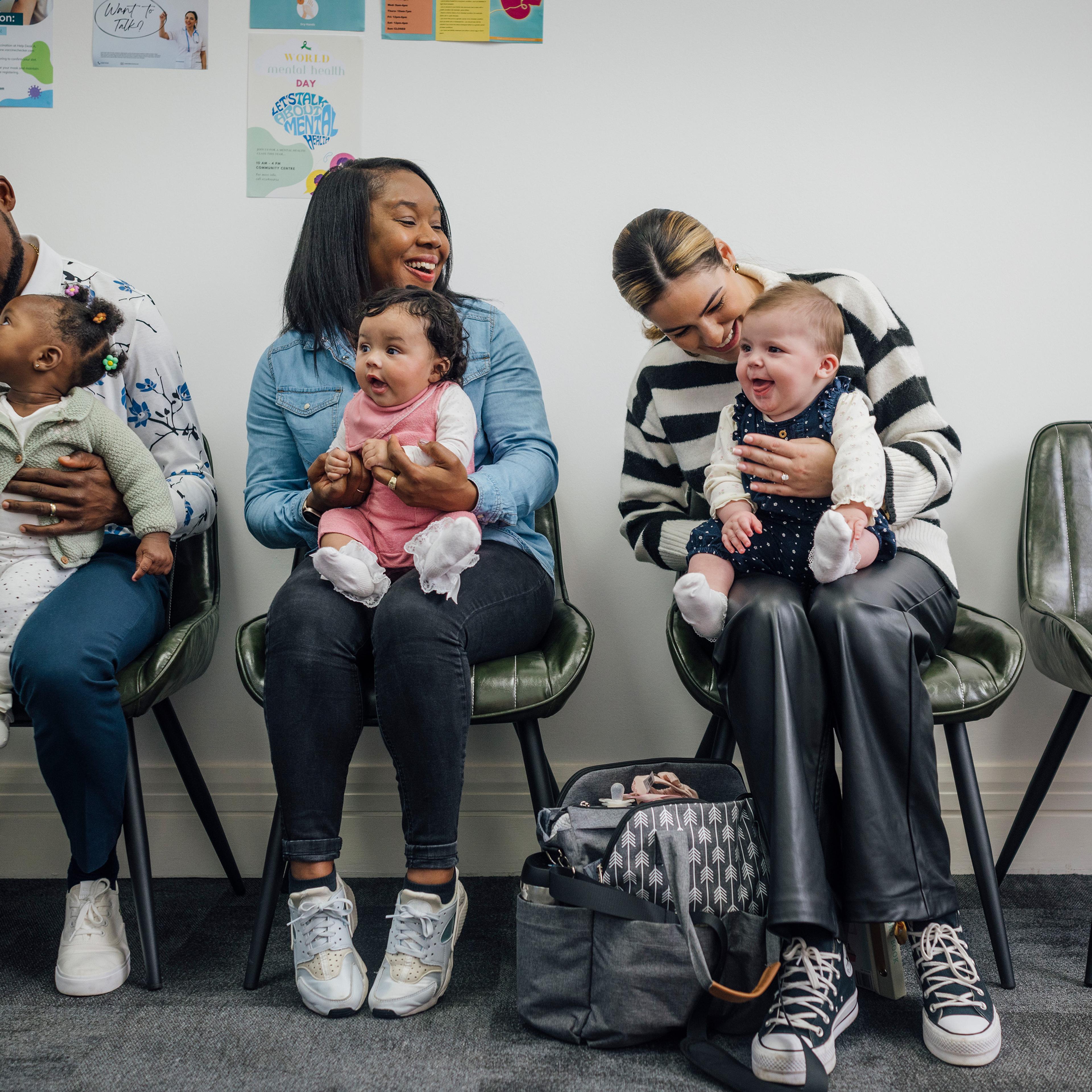 Three adults with babies seated in a waiting room, smiling and interacting, with colourful posters on the wall.