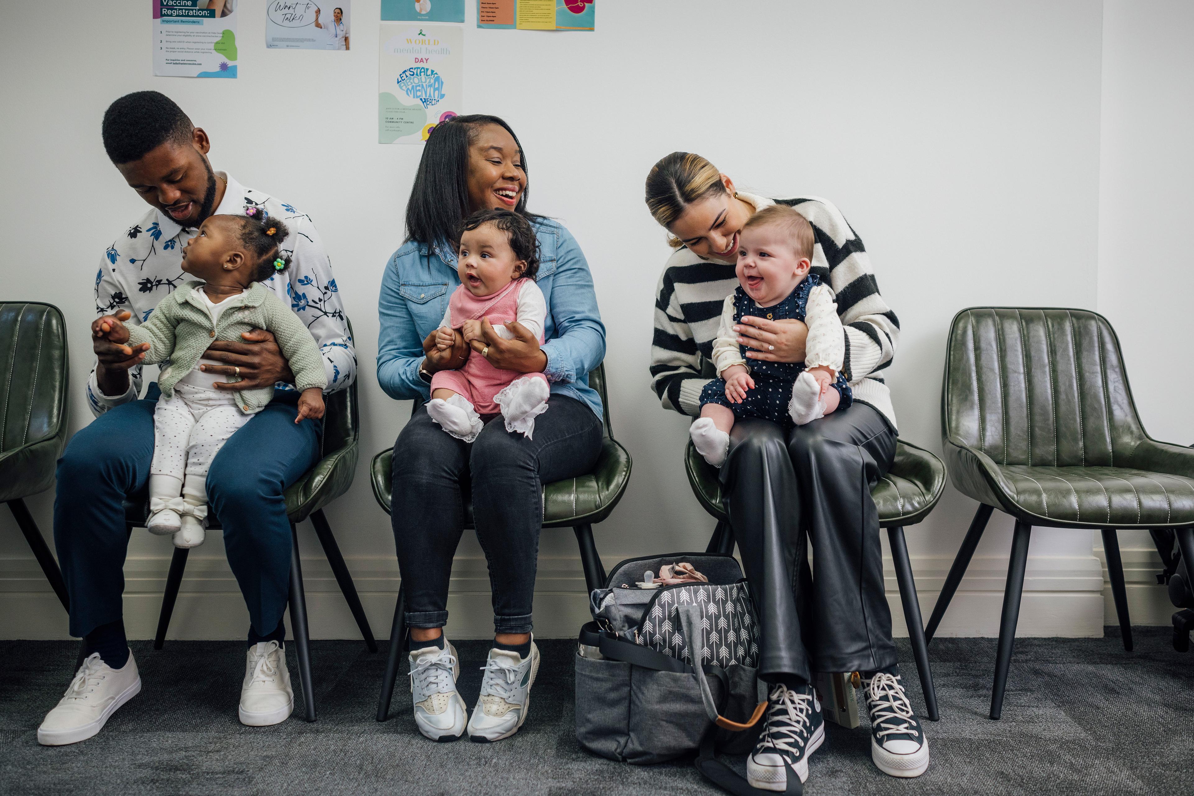 Three adults with babies seated in a waiting room, smiling and interacting, with colourful posters on the wall.