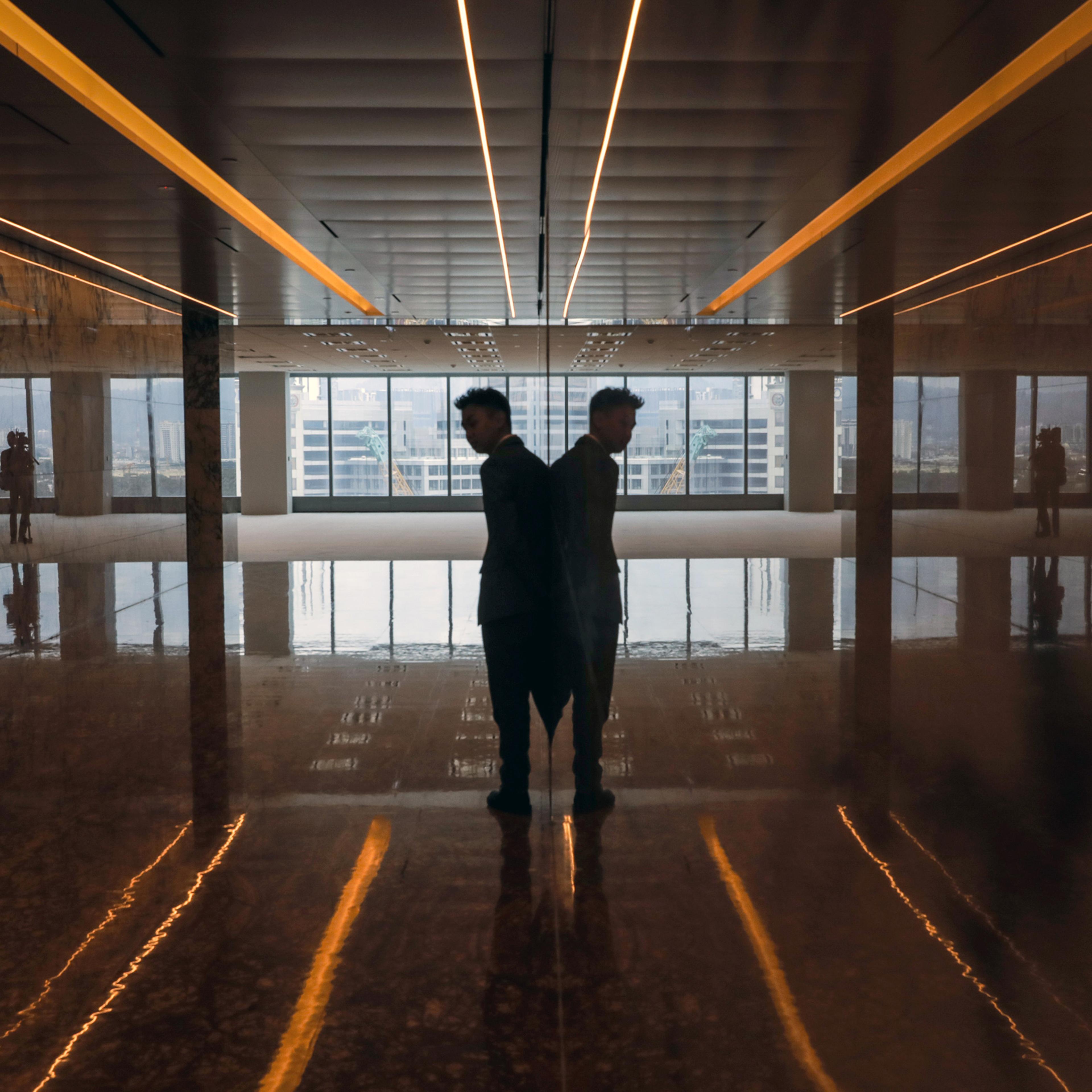 A man in silhouette standing against a reflective surface in an empty room with cityscape visible through large windows.