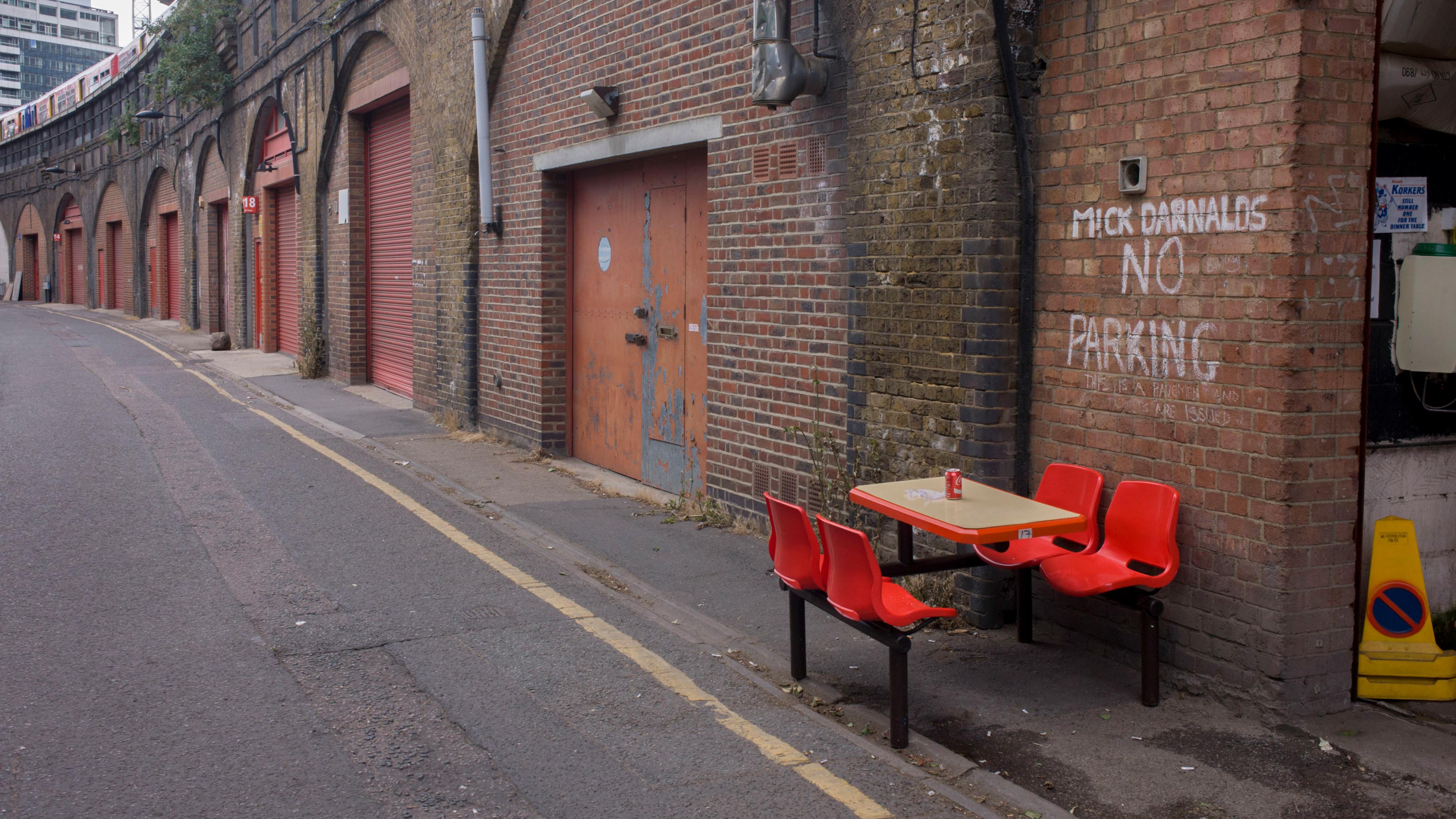A road below a railway line with a brick wall and red garage doors, graffiti that reads ‘MICK DARNALDS NO PARKING’ behind a table with a can of coke on it and four red chairs.