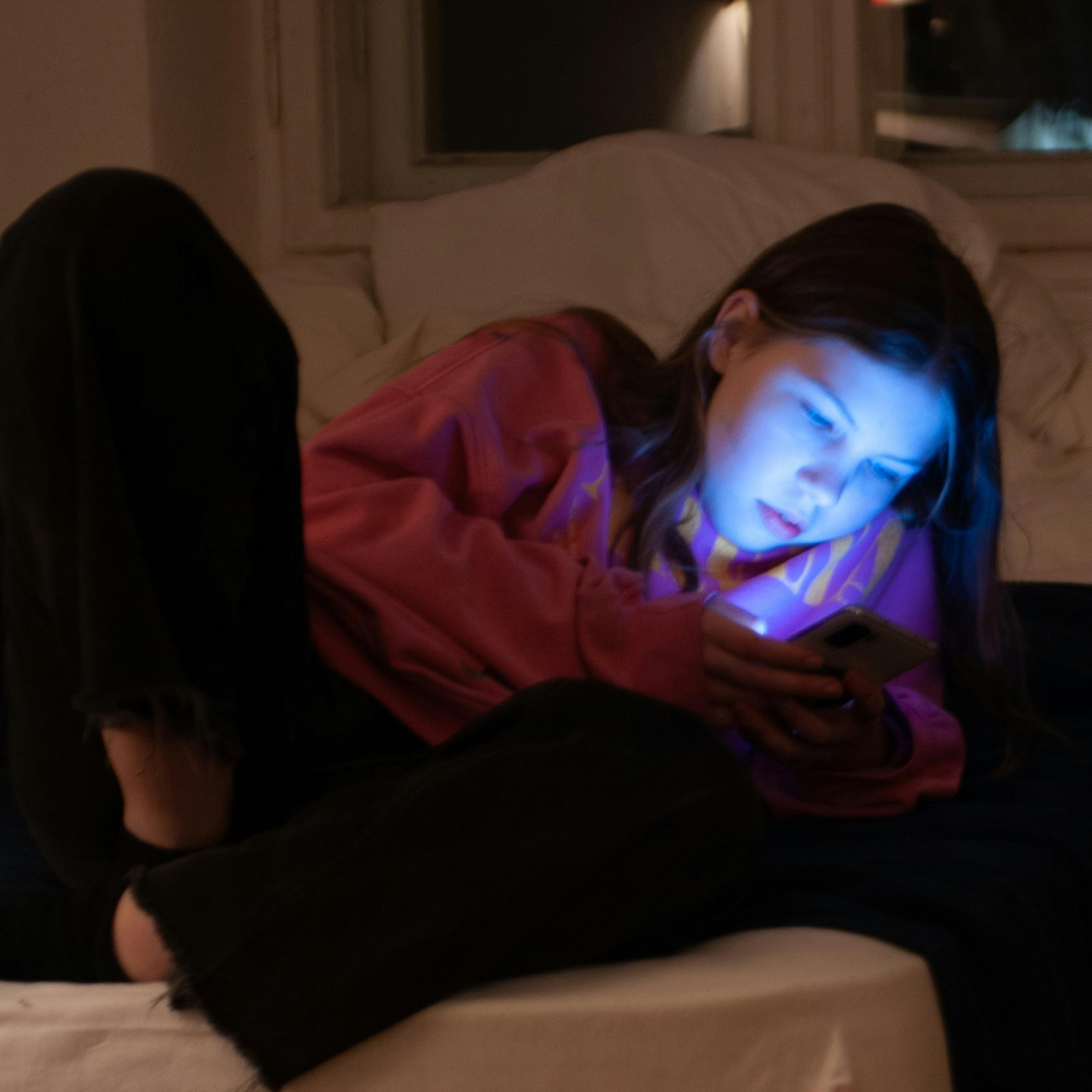 A young girl in a pink hoodie lit by a phone screen, lying on a bed in a dimly lit room at night.