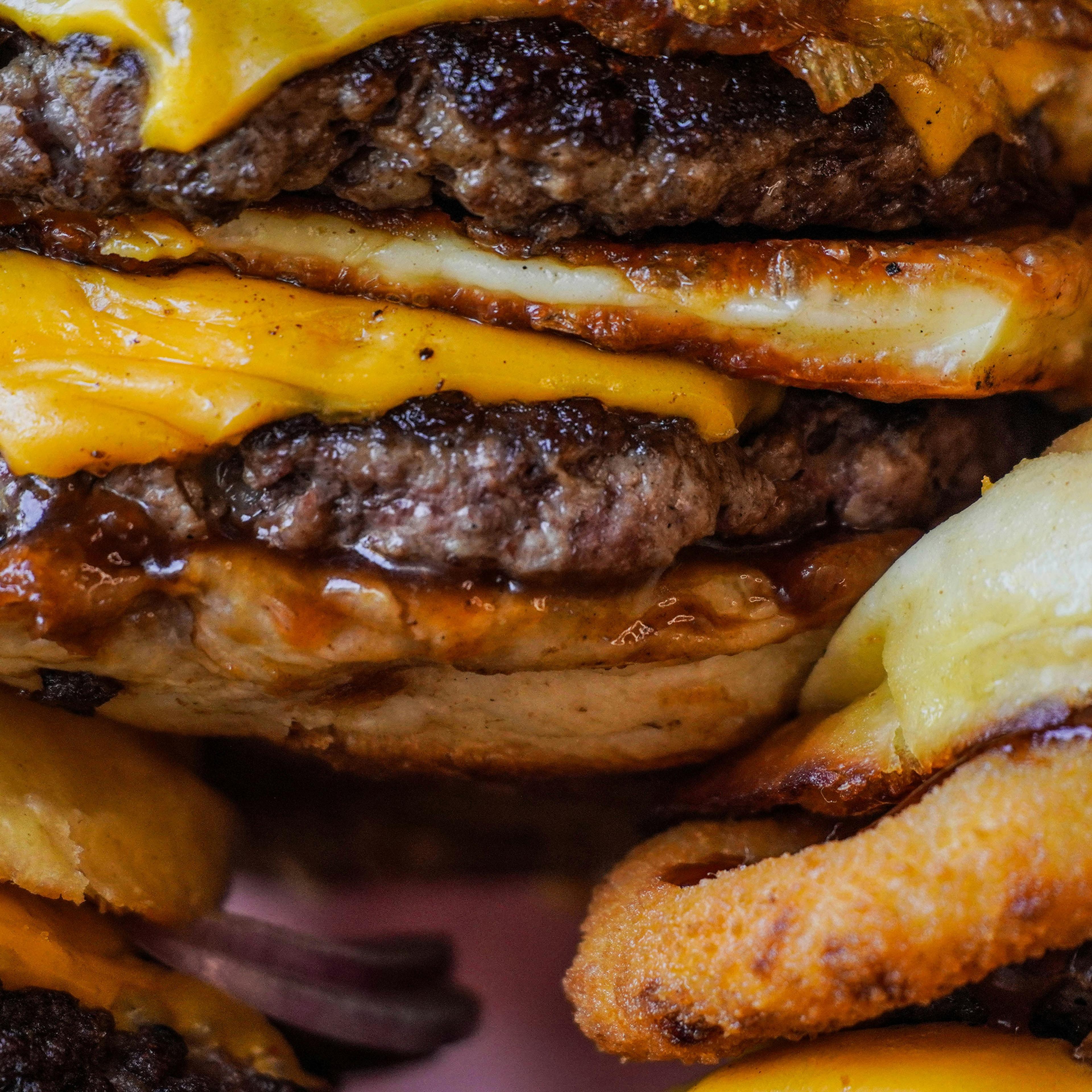 A close-up view of a cheeseburger with multiple beef patties, melted cheese and crispy onion rings.