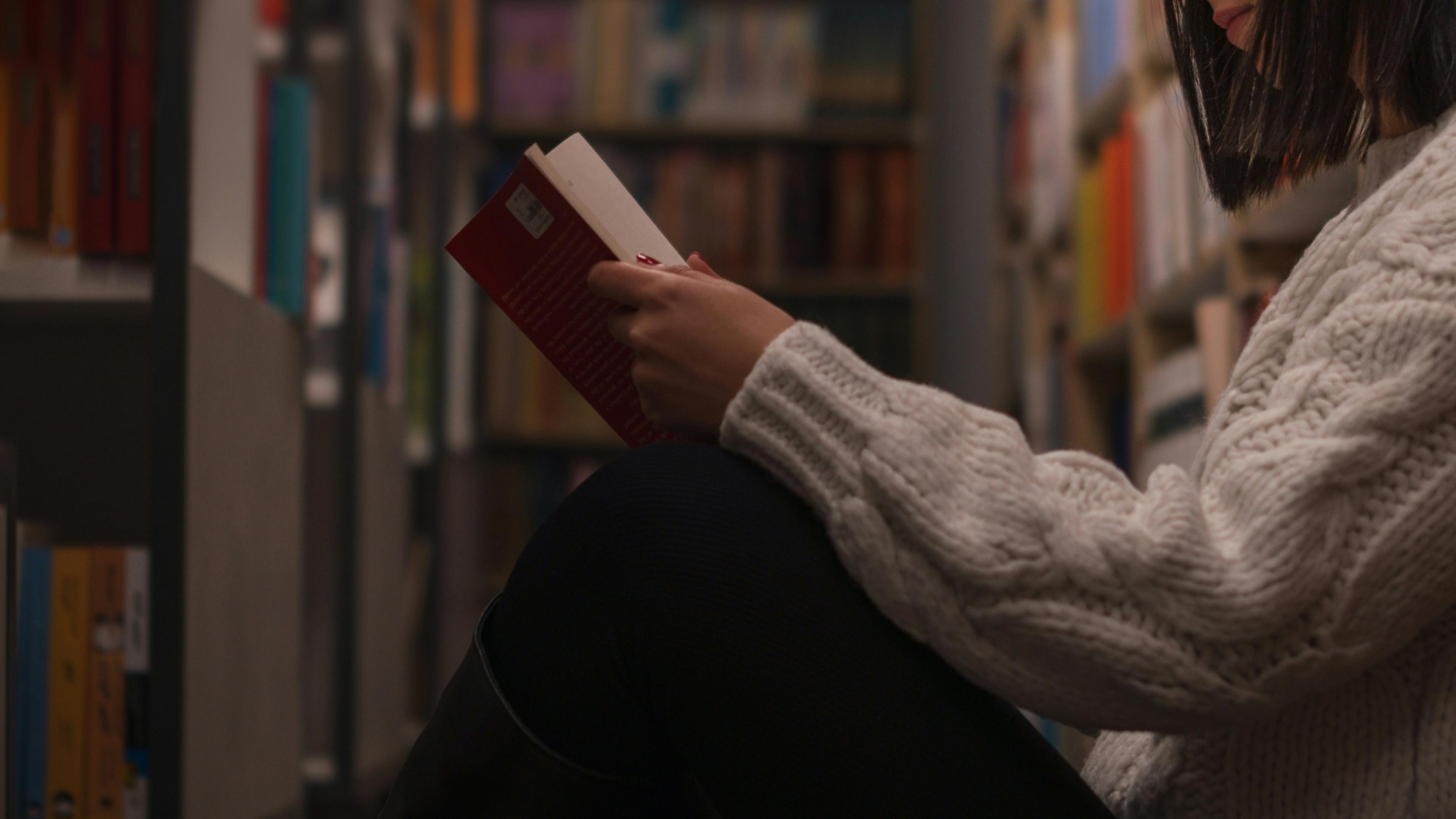 A person in a library sitting on the floor reading a red book wearing a white knitted jumper.