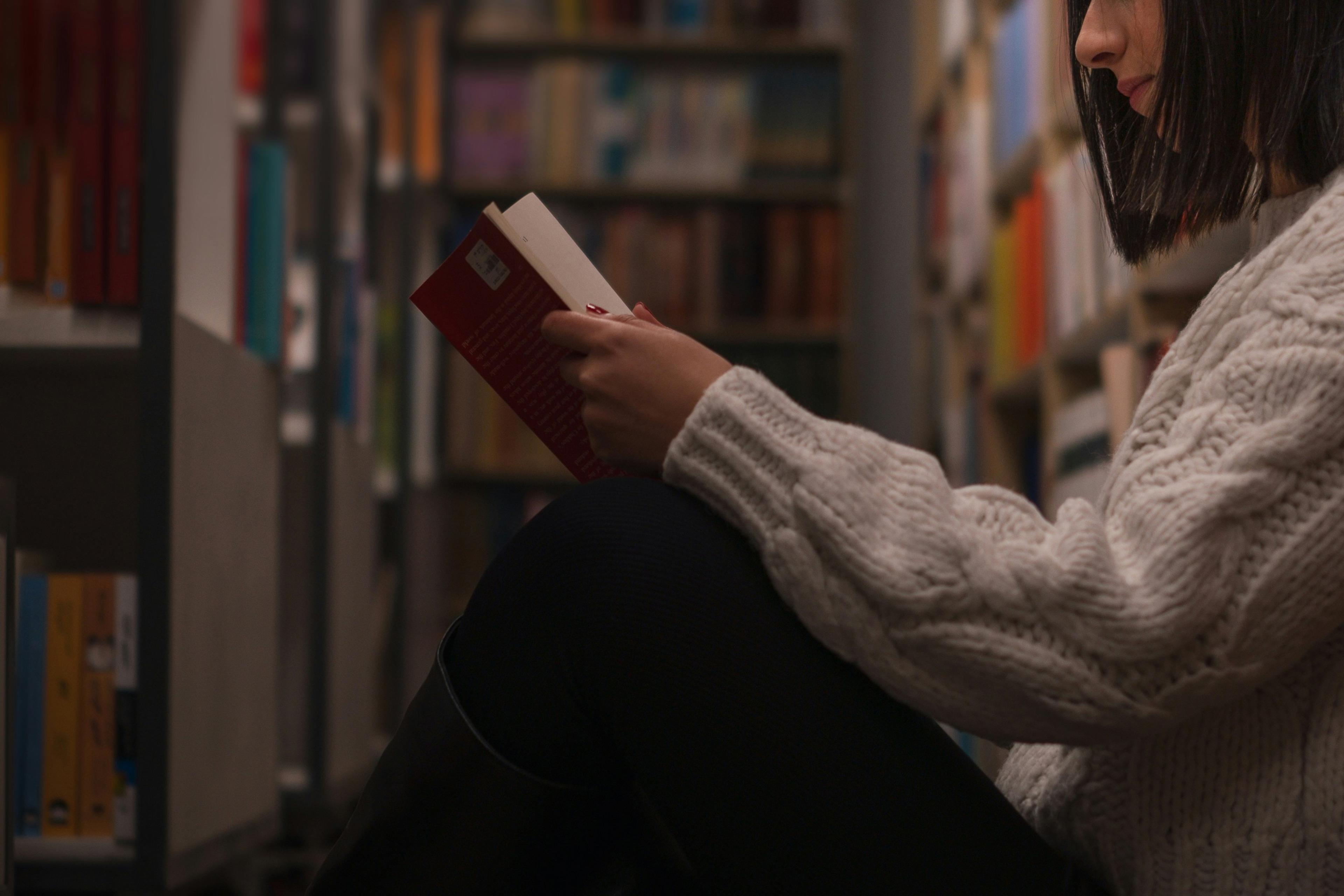 A person in a library sitting on the floor reading a red book wearing a white knitted jumper.