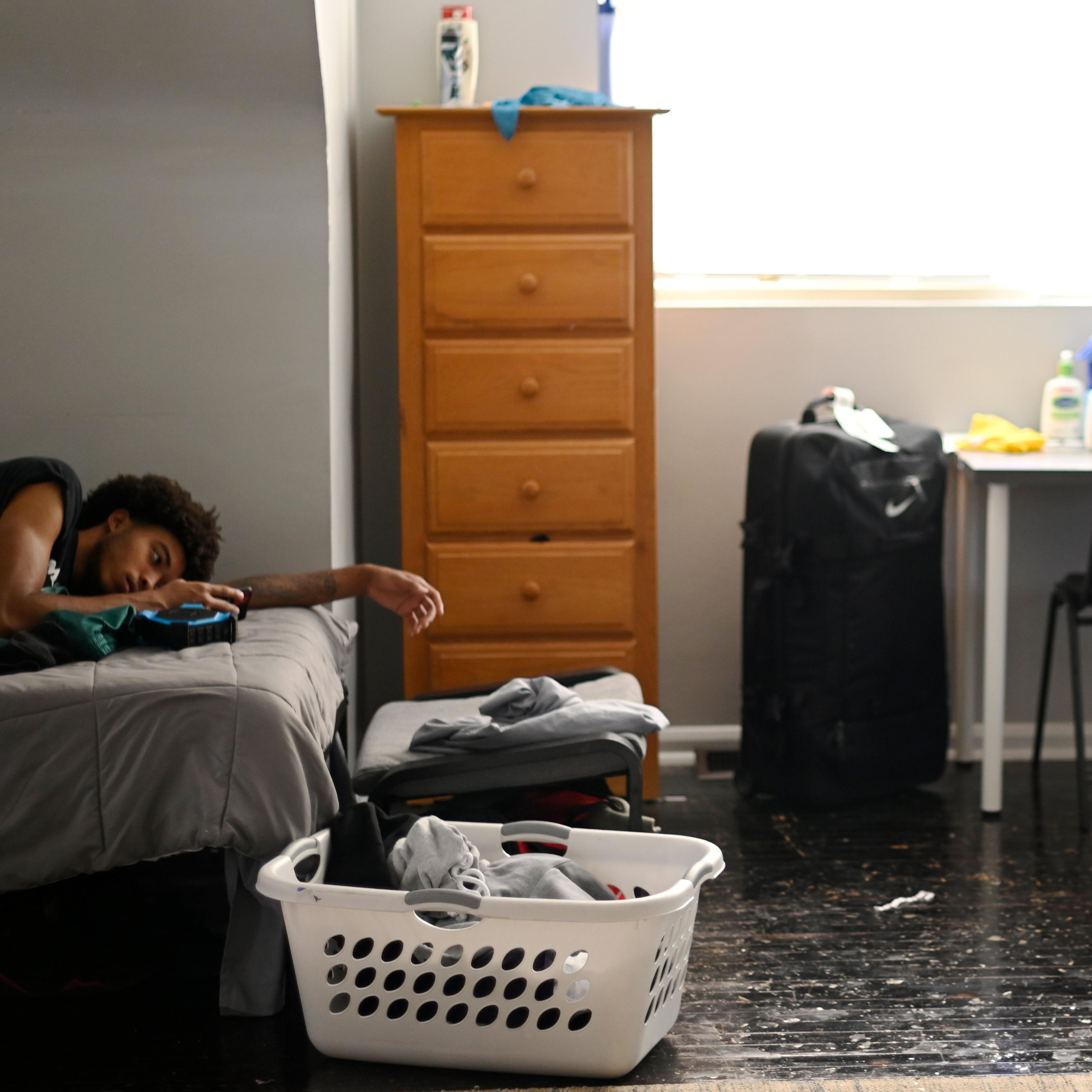 A young person lying on a bed in a cluttered room with a suitcase, laundry basket, chest of drawers and desk, illuminated by window.