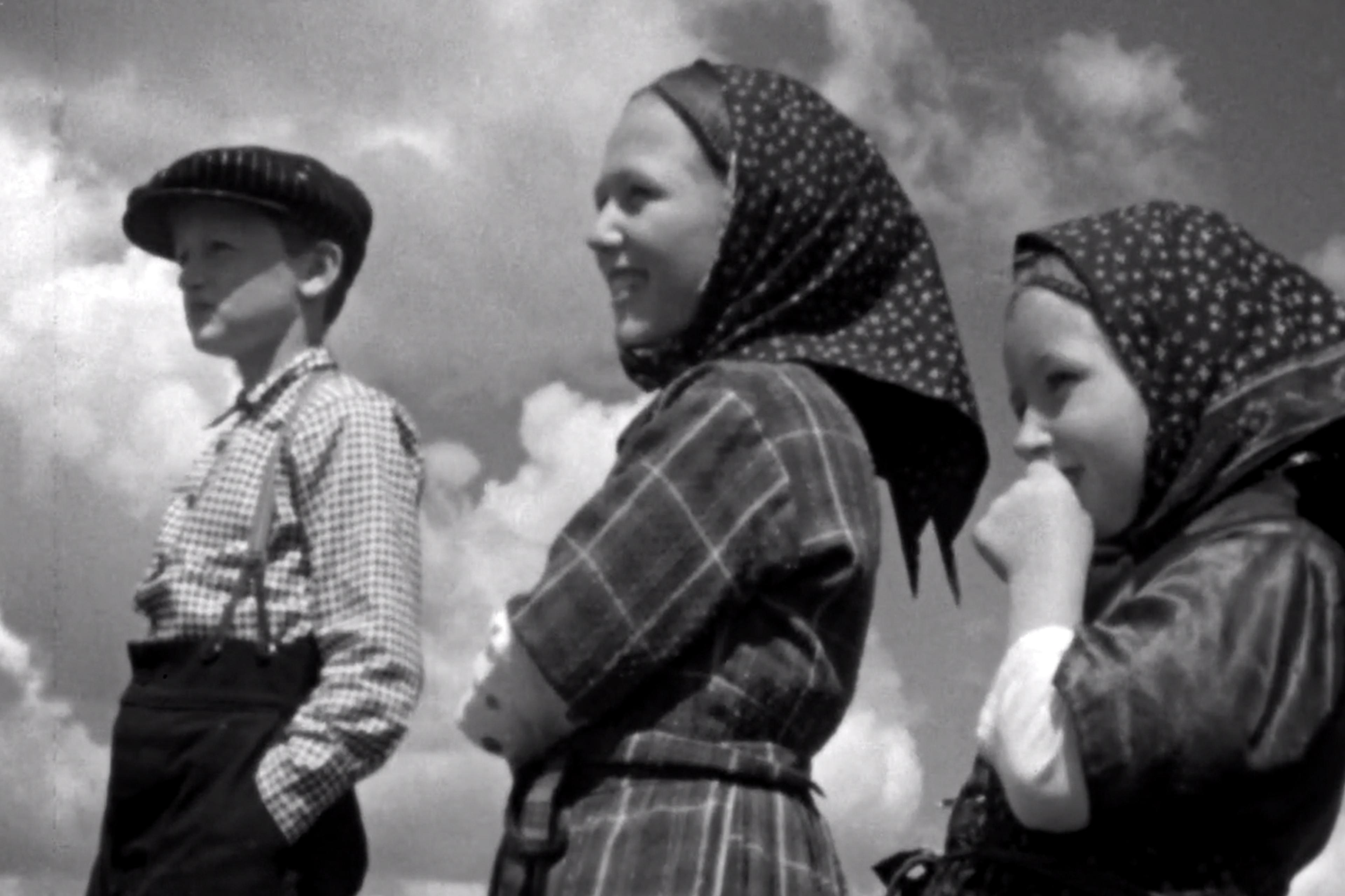 Black and white photo of three children standing outdoors under a cloudy sky. The boy on the left wears a flat cap and checked shirt with trousers, while the two girls on the right wear patterned headscarves and dresses, smiling and looking into the distance.