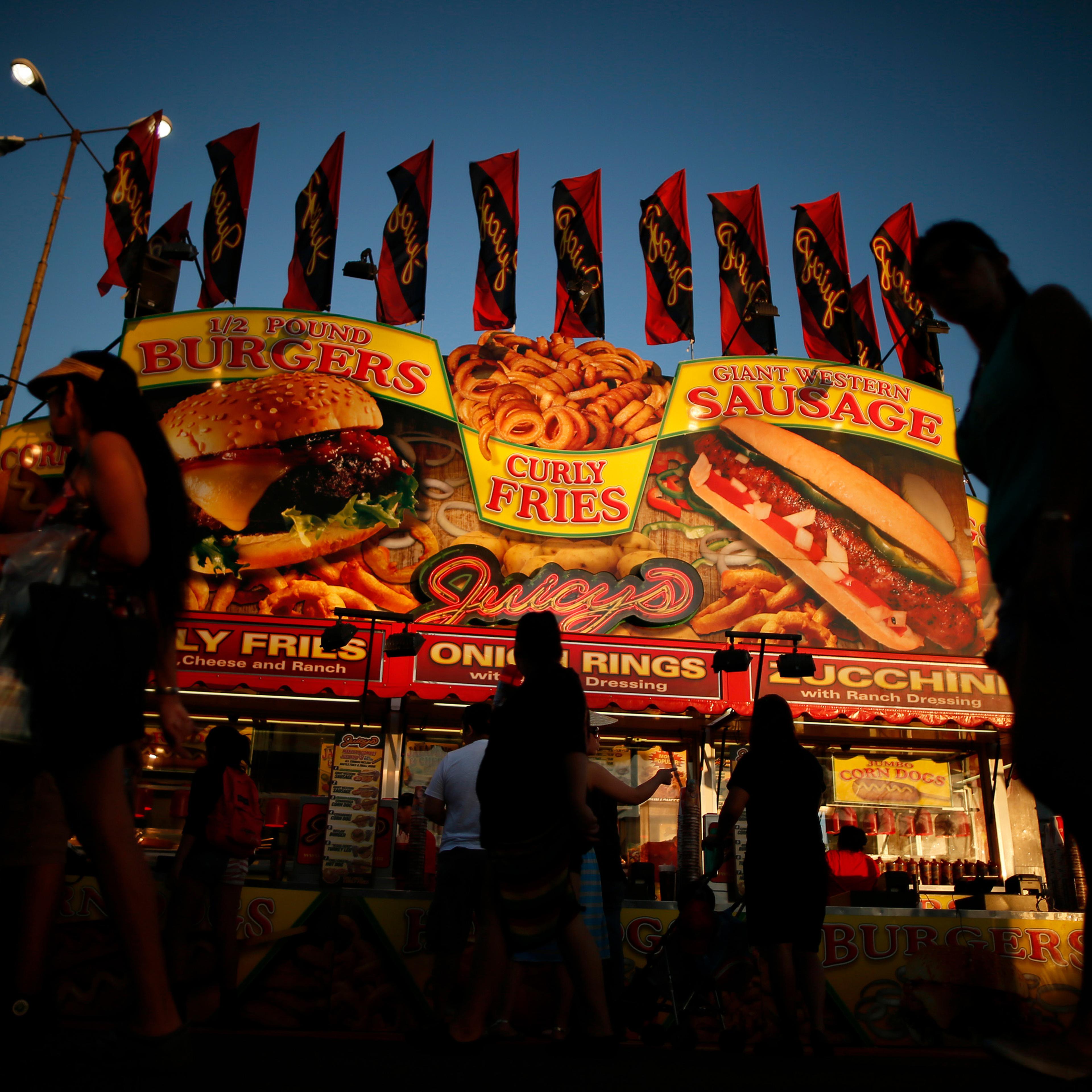 A bustling food stall at dusk with bright signs advertising burgers, curly fries and sausages against a darkening sky.