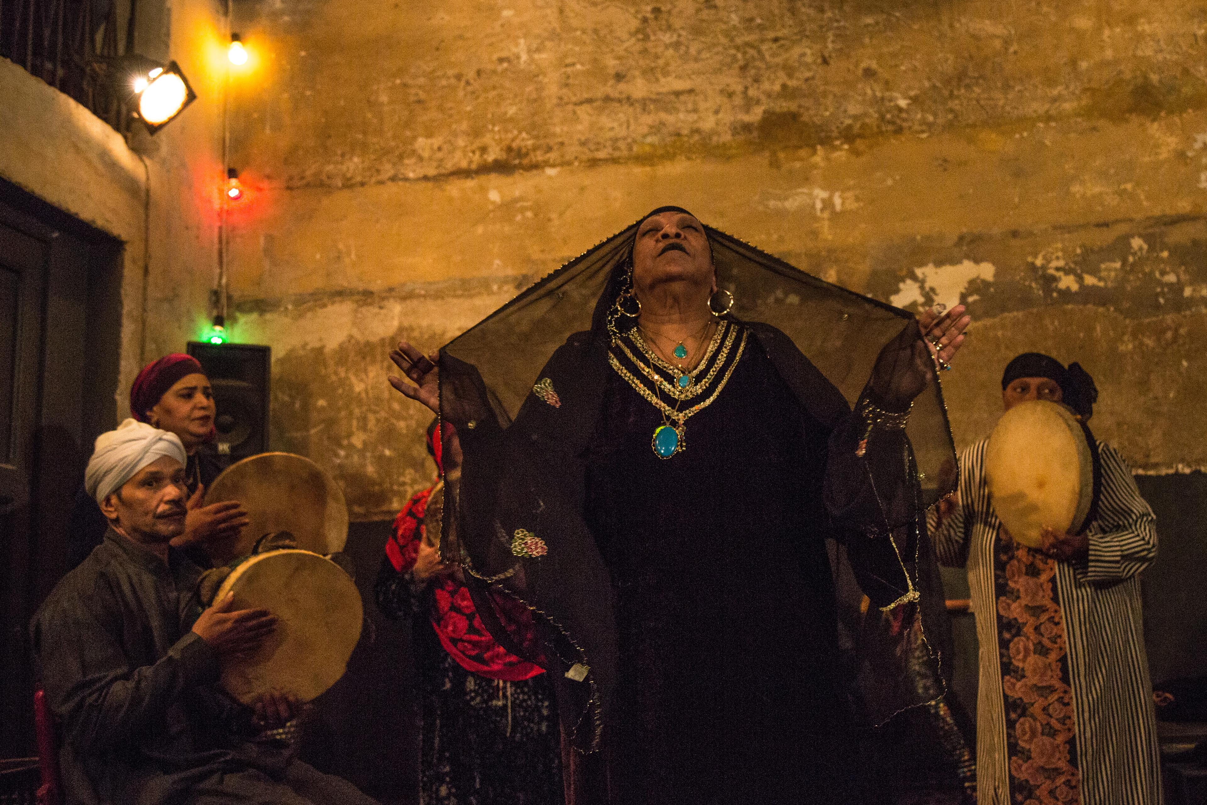 A woman wearing a black gown and veil, and gold and turquoise jewellery raises her harms with eyes closed; musicians wearing headdresses play drums in the background in a dimly lit setting.