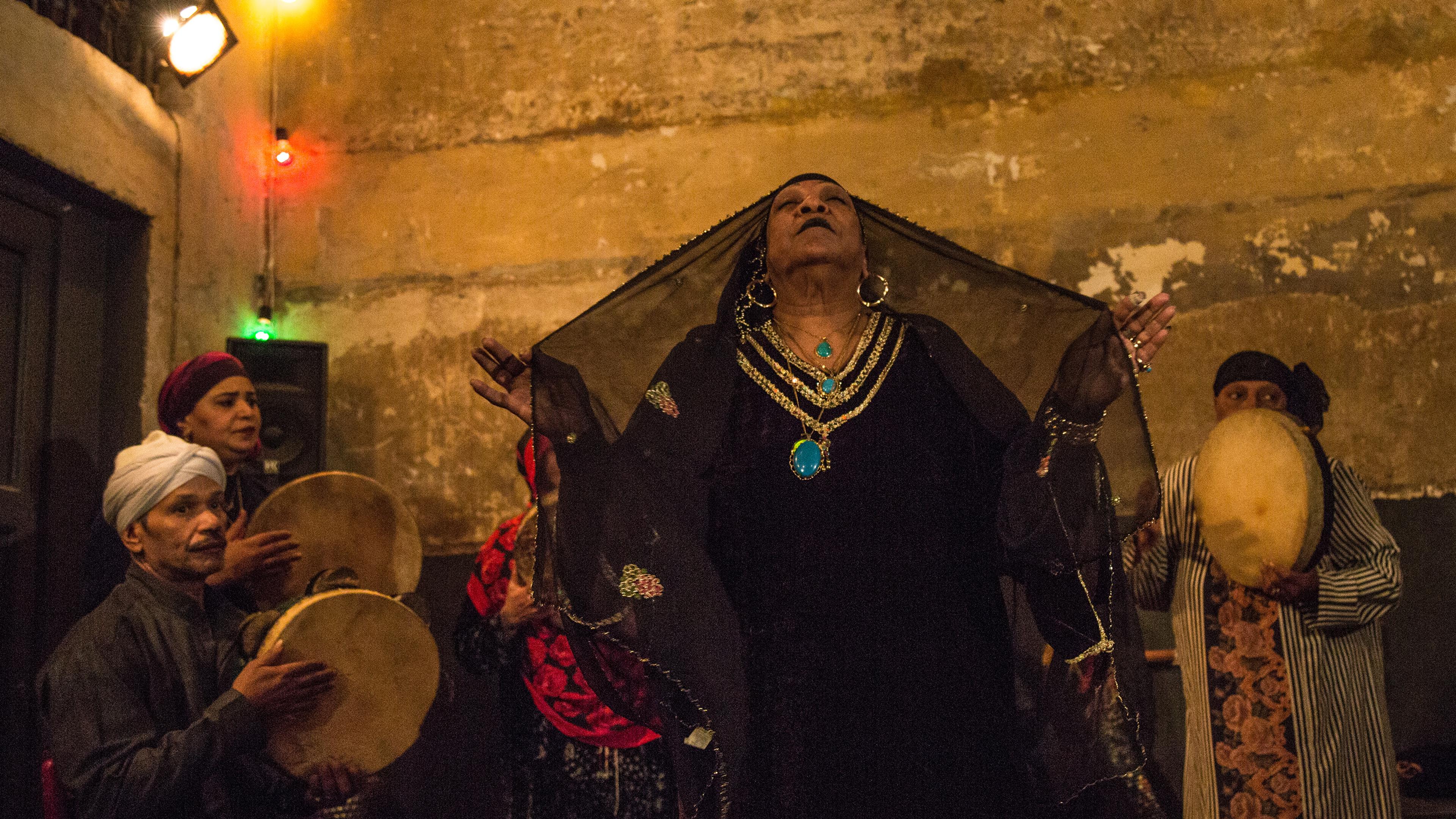 A woman wearing a black gown and veil, and gold and turquoise jewellery raises her harms with eyes closed; musicians wearing headdresses play drums in the background in a dimly lit setting.
