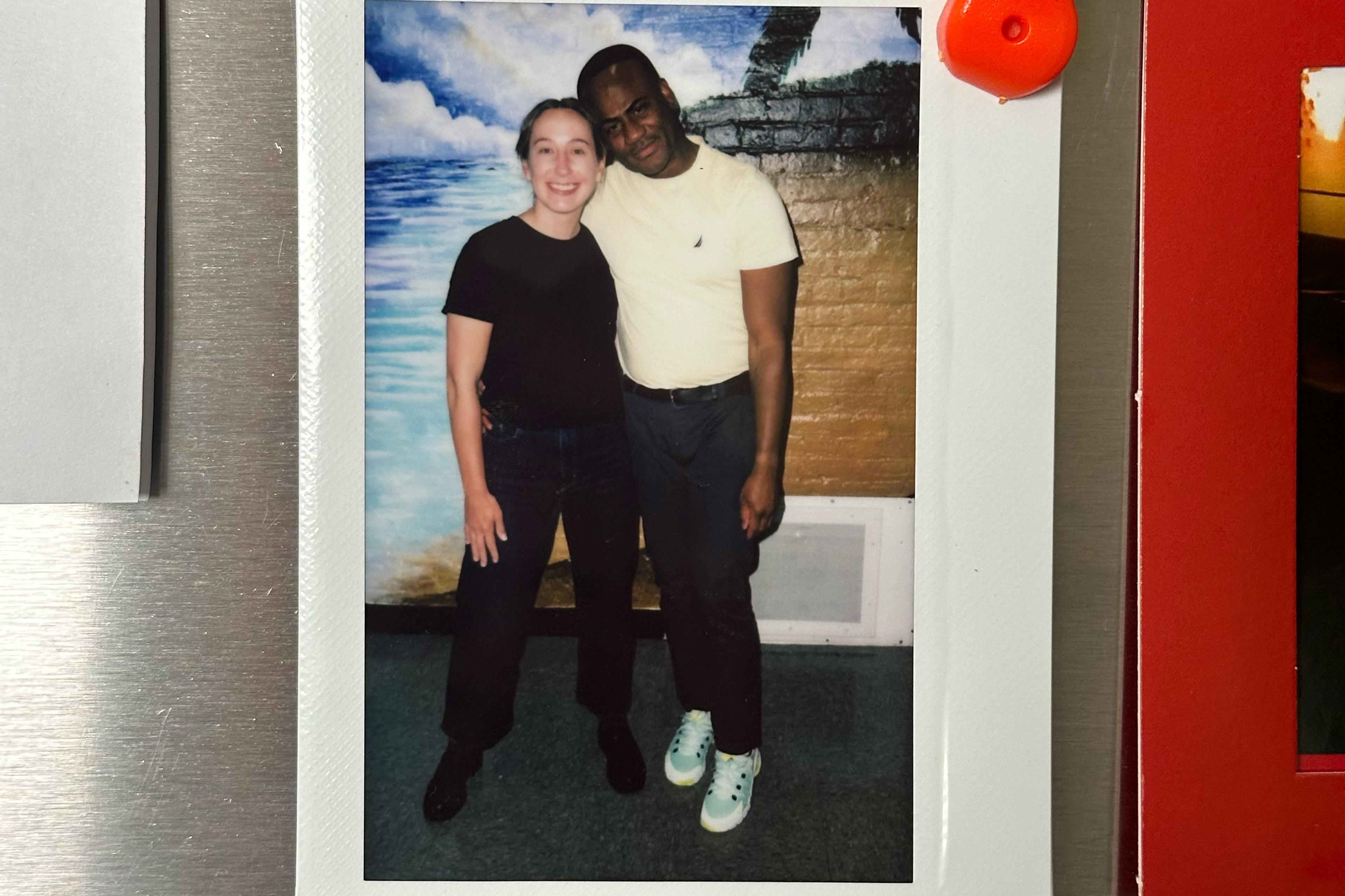 A Polaroid photo of two people standing together smiling in front of a painted mural depicting the sea and clouds.