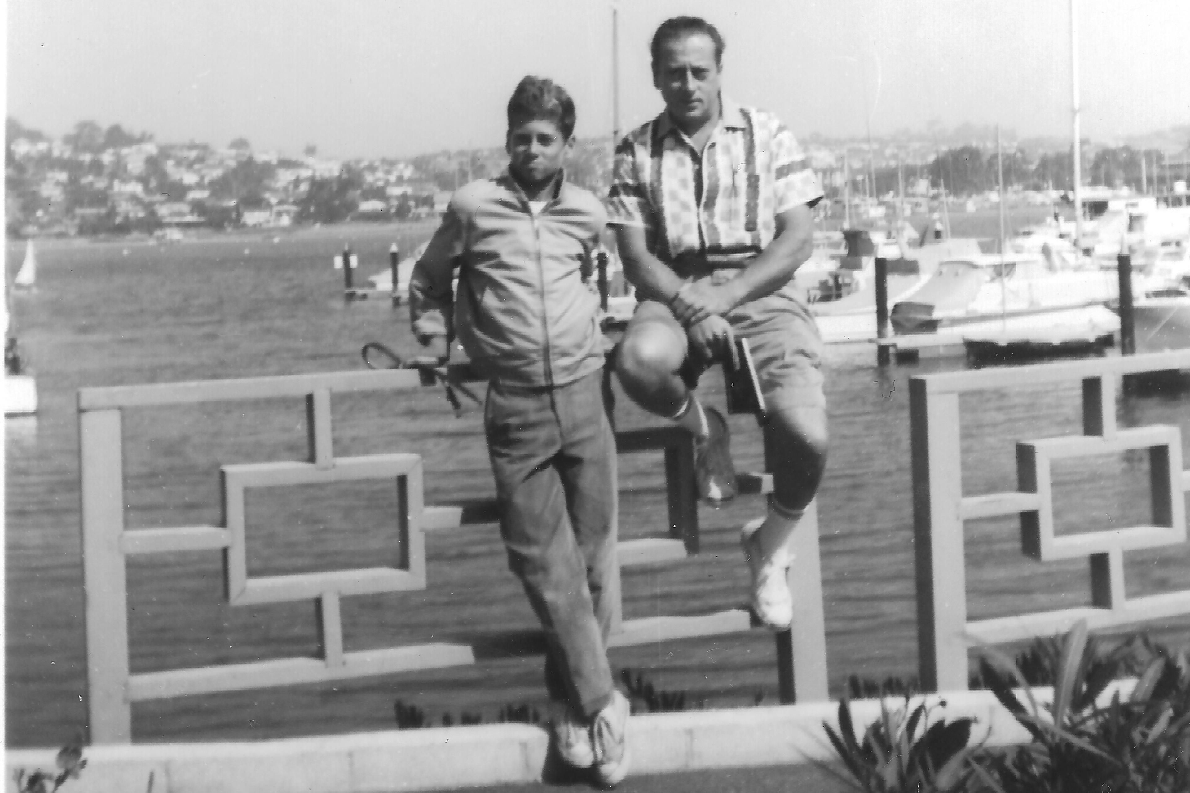 Black and white photo of two men by a waterfront fence, with boats and a hillside town in the background.