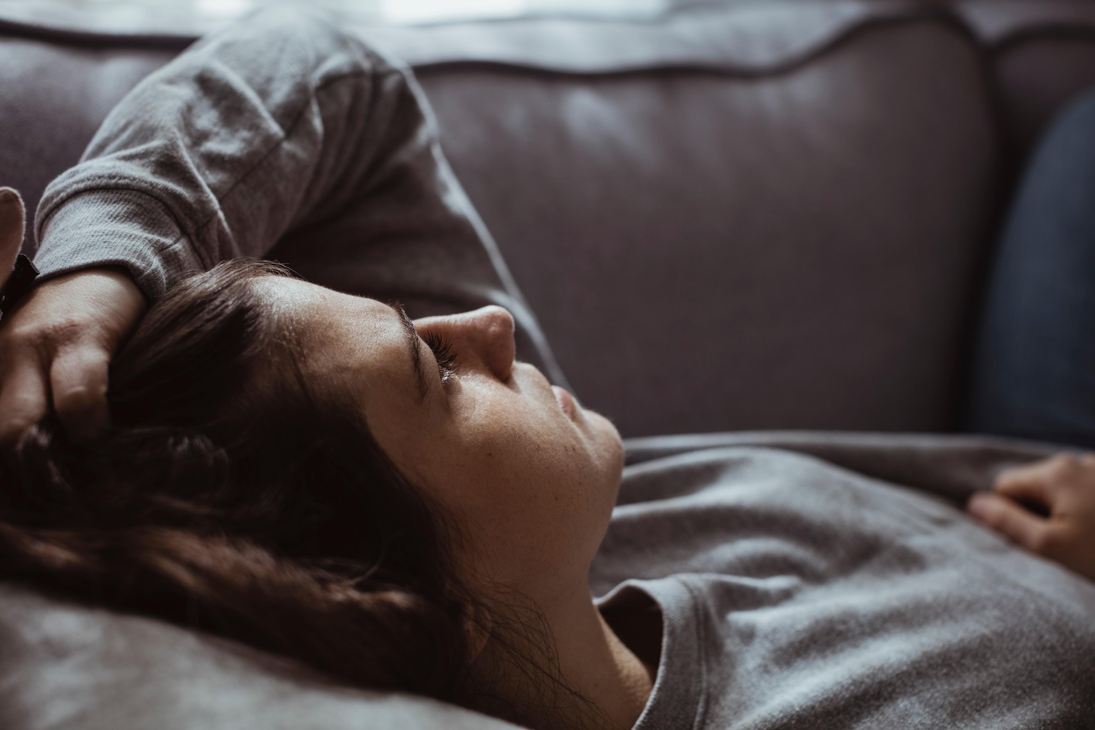 Photo of a person lying on a sofa, looking relaxed with eyes closed. They are wearing a grey jumper and holding their hair.