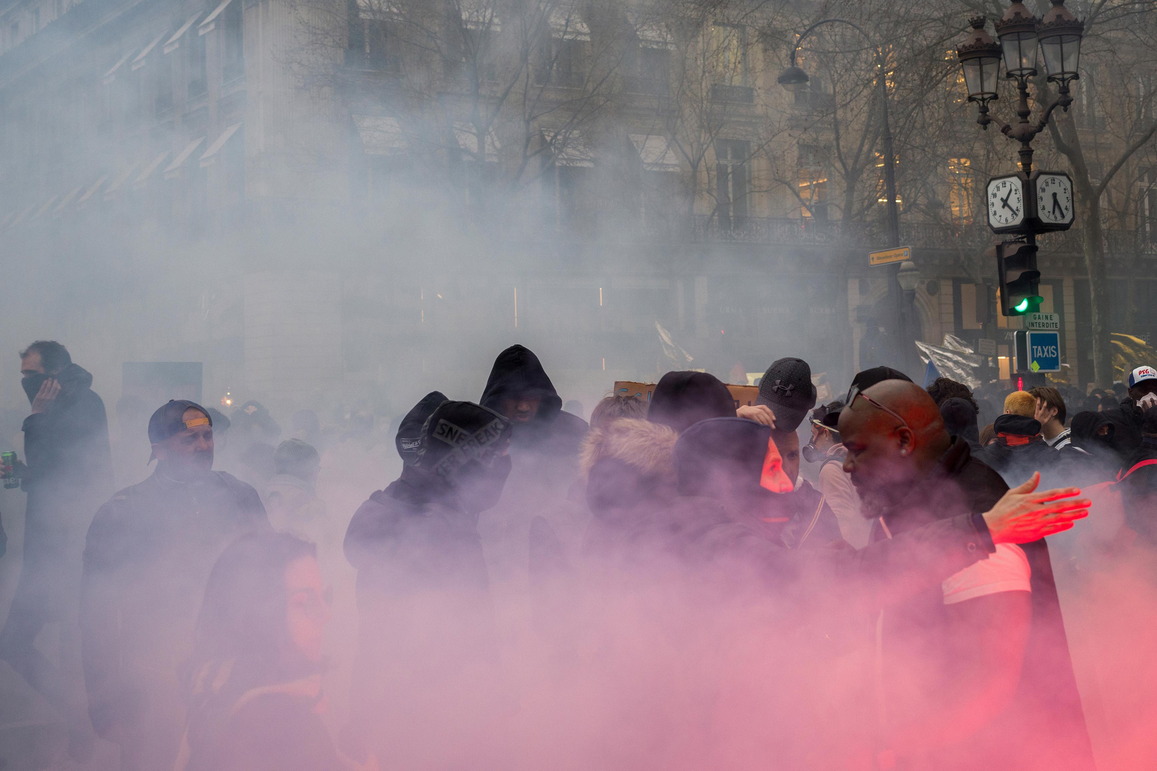 A crowd enveloped in pink smoke on a city street, with people wearing hoodies and jackets, and buildings in the background.