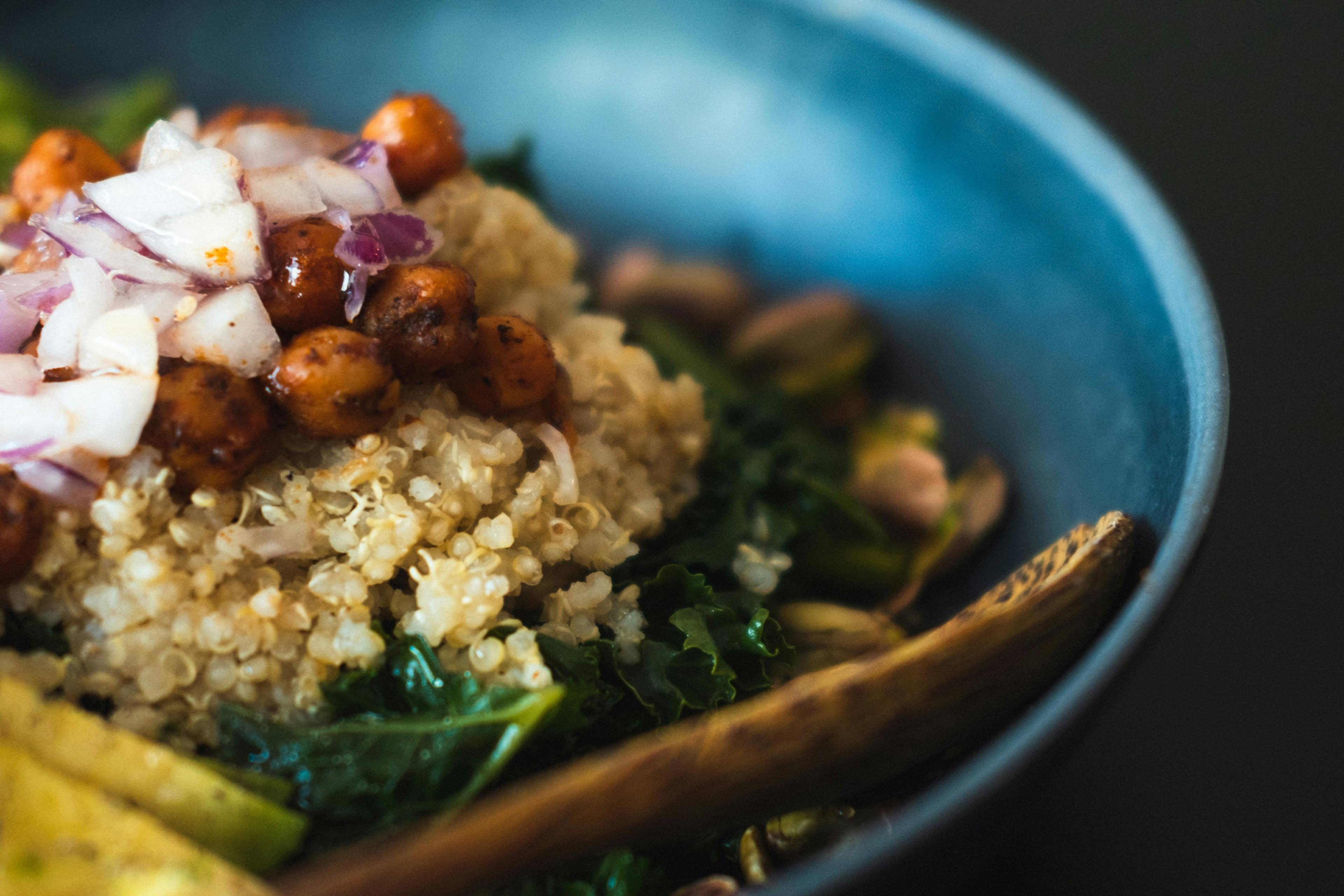 A blue bowl with quinoa, chickpeas, chopped onions, greens and a wooden spoon on a dark background.