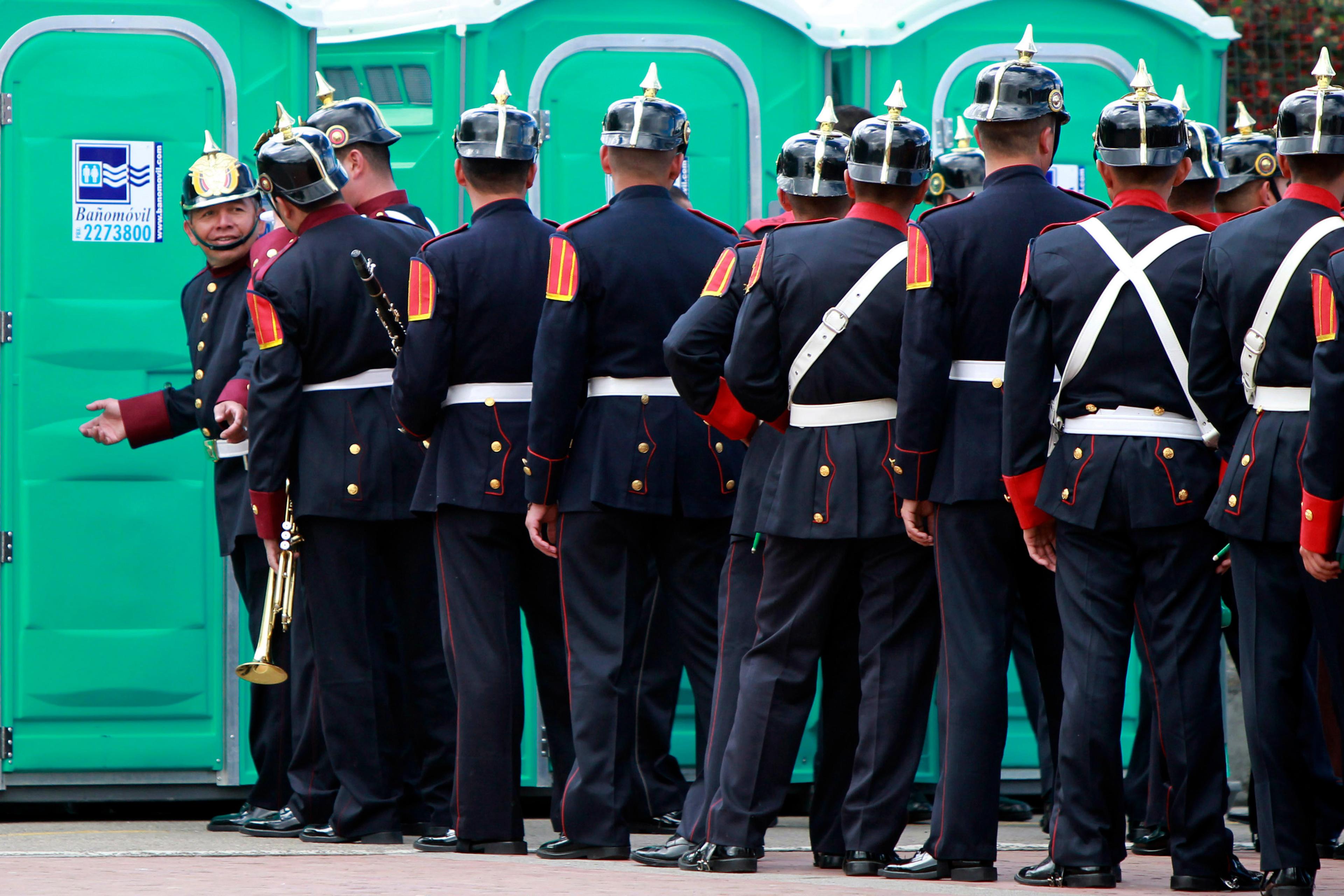 Photo of uniformed men standing outside portable toilets, one gestures invitingly.