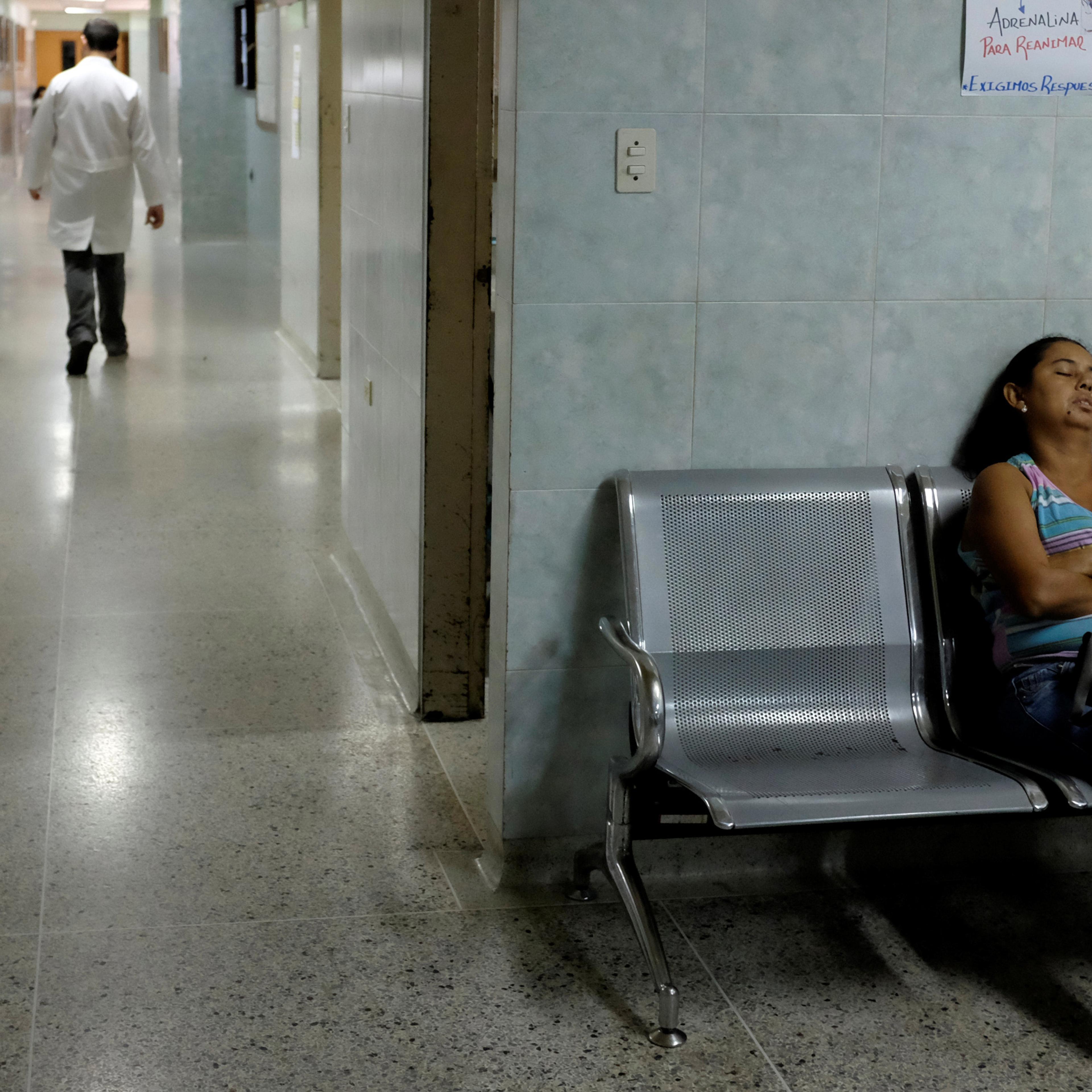 A hospital corridor with a woman asleep on a bench and a doctor walking away in the background.
