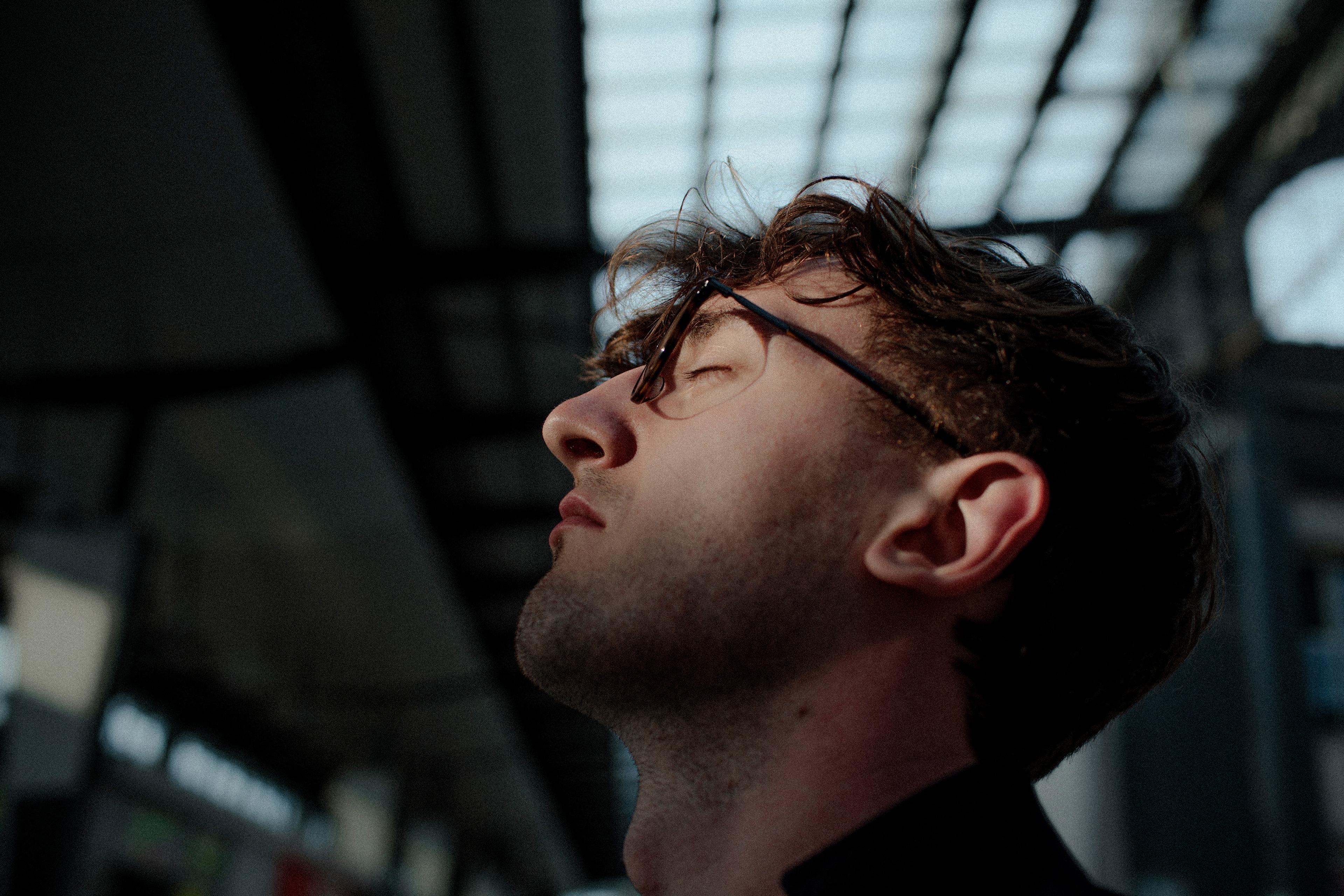 Photo of a man in profile with glasses, eyes closed, head tilted back under a structure with a grid-like roof.