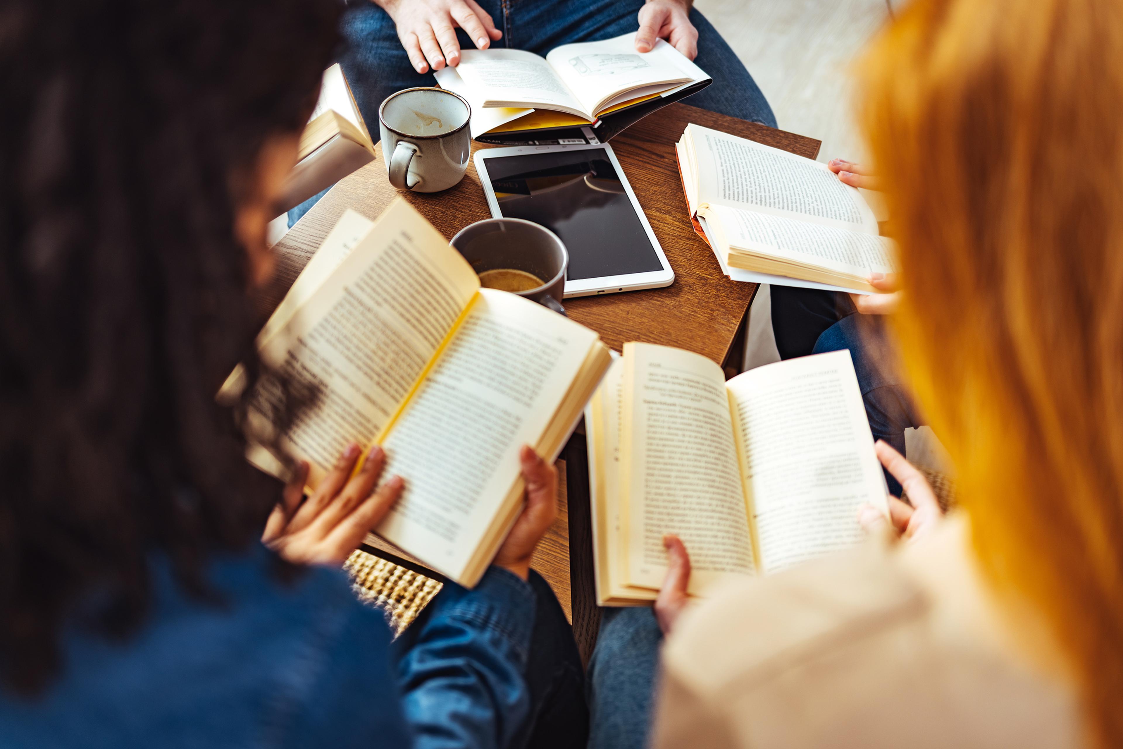 People sitting around a table reading books with a tablet and mugs visible.