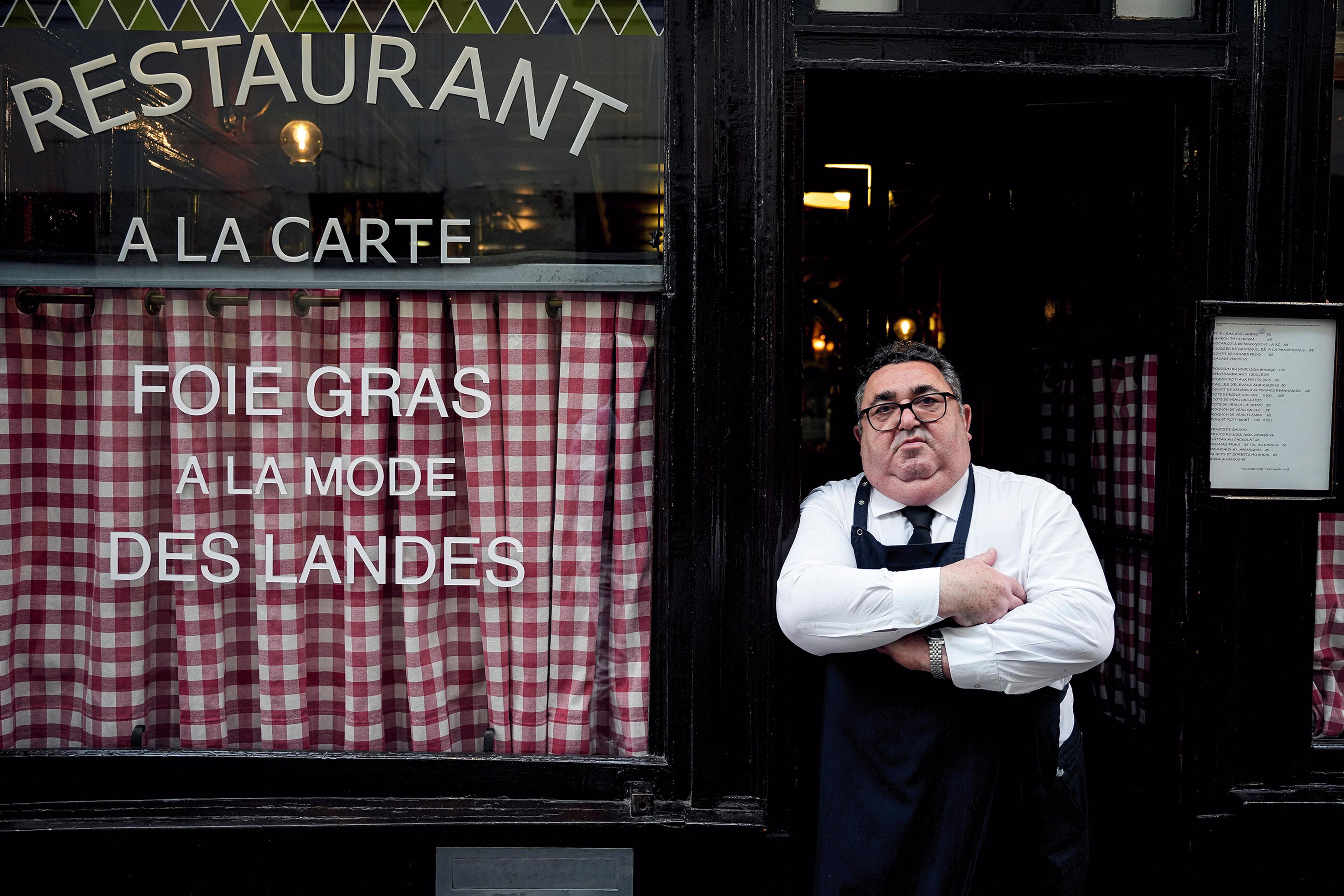 A man with crossed arms and a serious expression wearing an apron stands in the doorway of a restaurant with checkered curtains and menu on display.