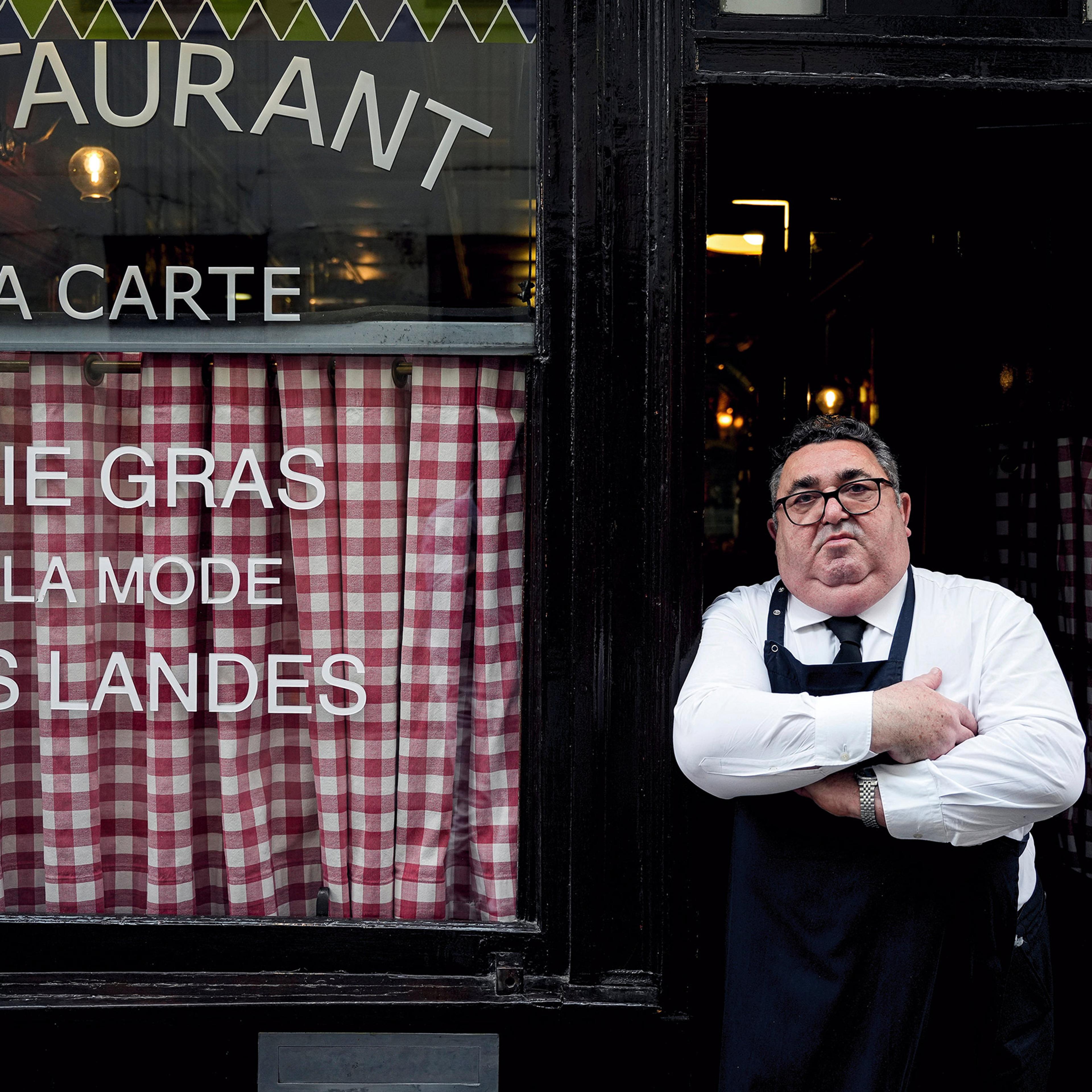 A man with crossed arms and a serious expression wearing an apron stands in the doorway of a restaurant with checkered curtains and menu on display.