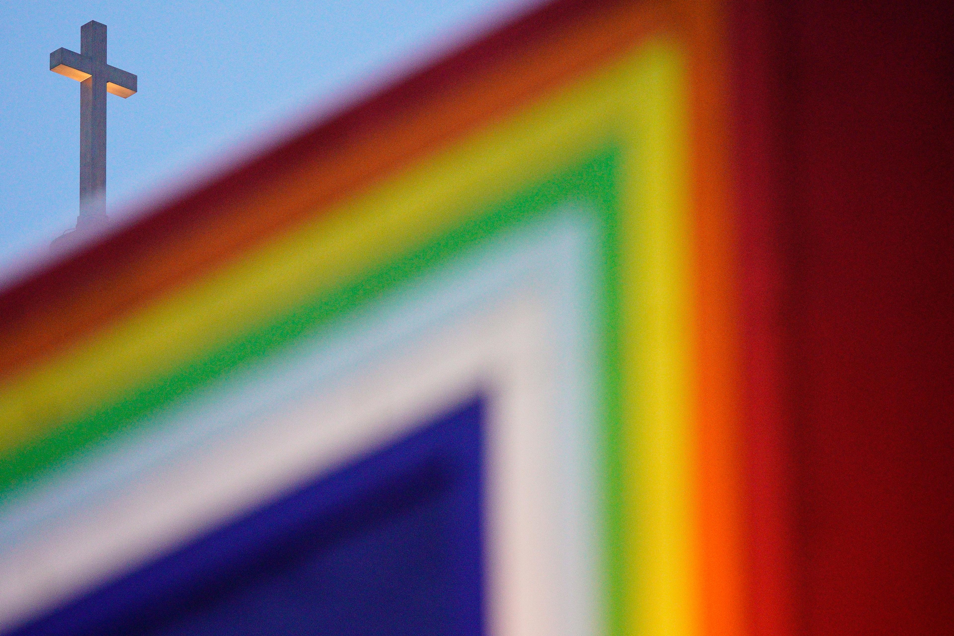 Photo of a cross in the sky with a blurred rainbow-coloured flag in the foreground.