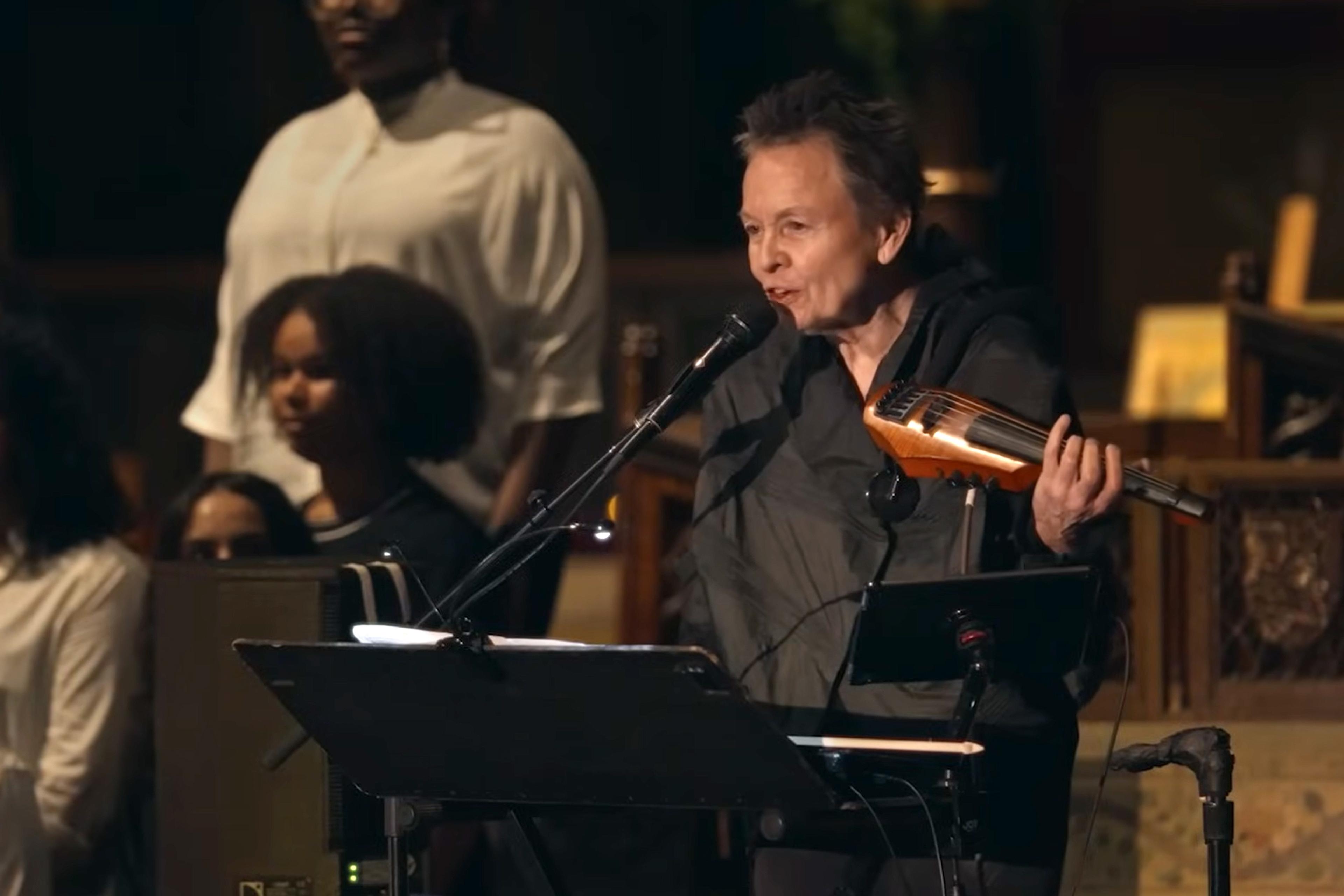 Photo of a person speaking into a microphone holding an electric string instrument with a choir in the background.